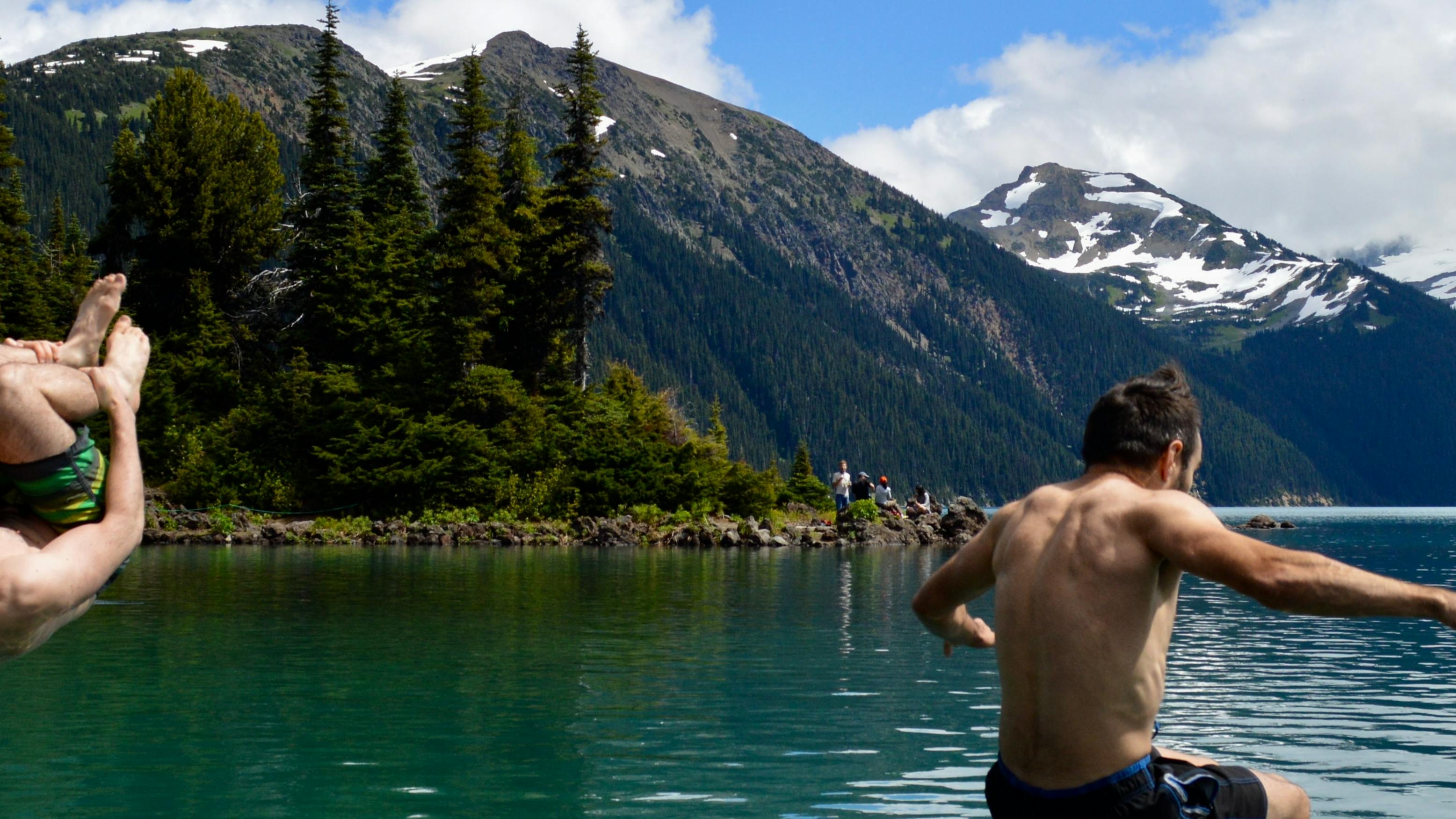 Two men jump into a lake. There are snow-topped mountains in the background. 