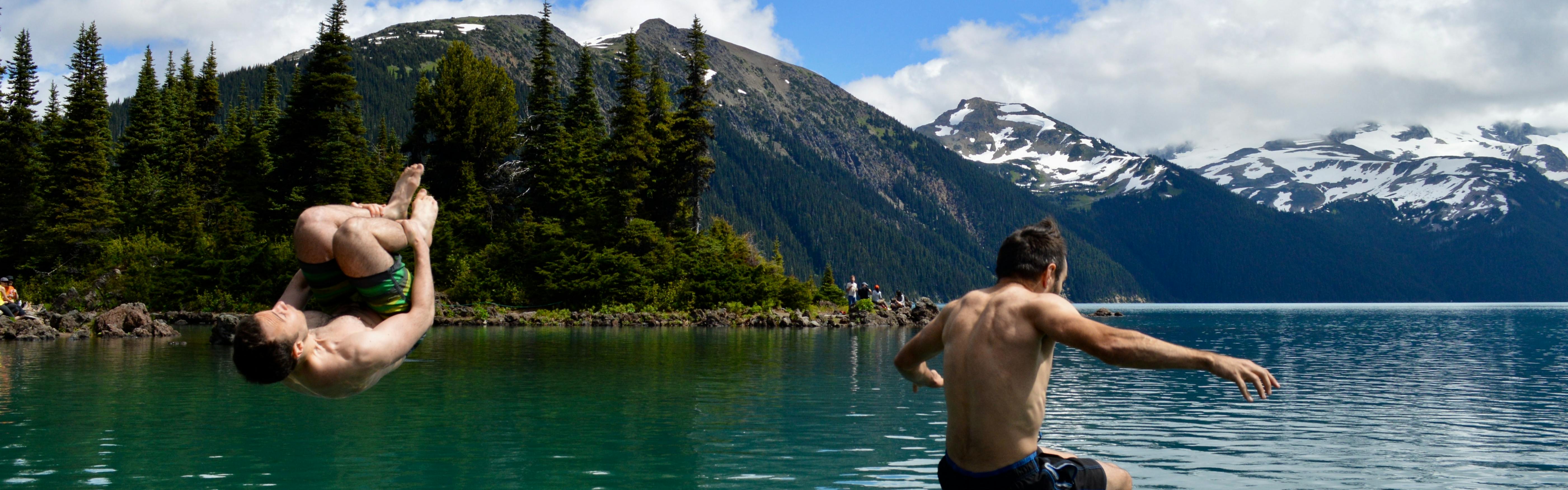 Two men jump into a lake. There are snow-topped mountains in the background. 