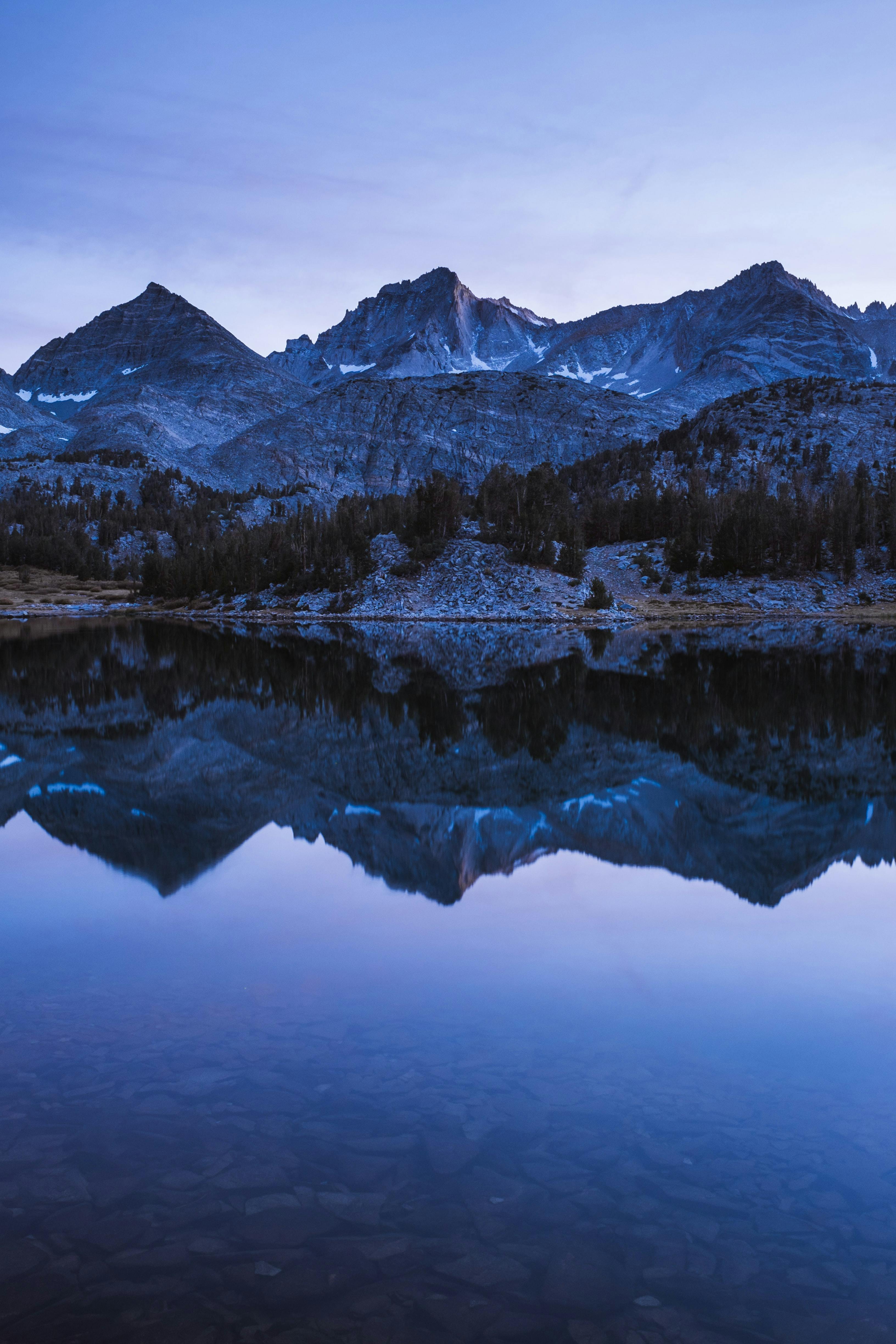 Mountains are reflected in clear, still water. The scene is dusky indigo. 