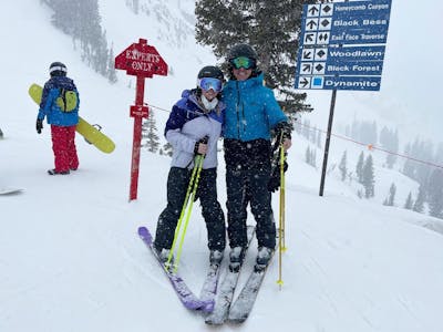 Two skiers stand on a ski run at a resort. One has his hand on the other. There are trail signs behind them.