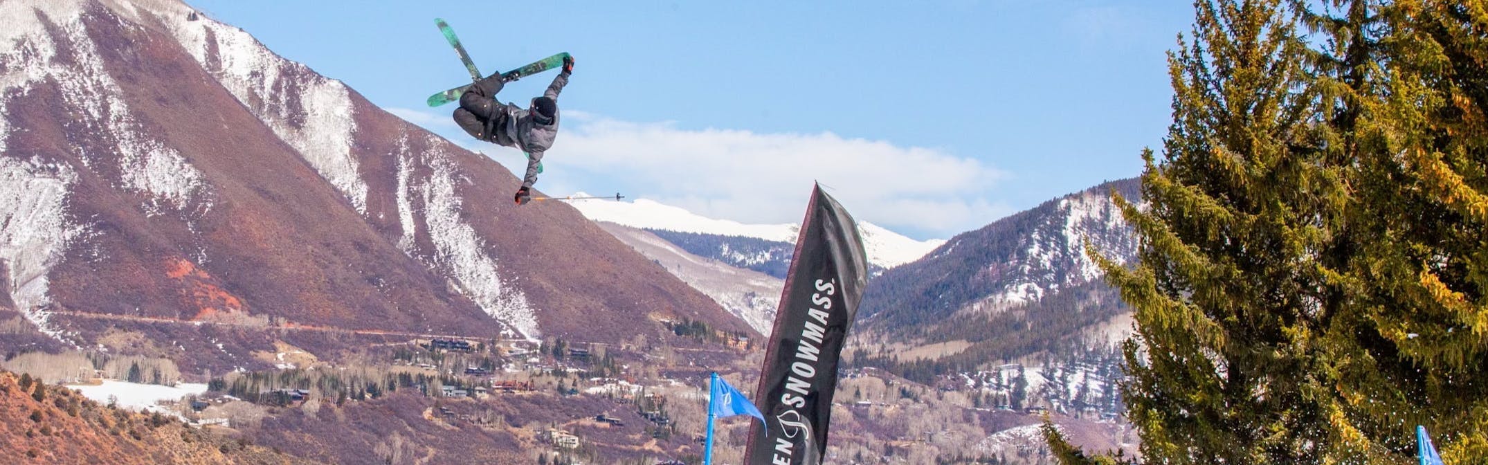 A skier goes off a jump at a ski resort. There are mountains in the background.