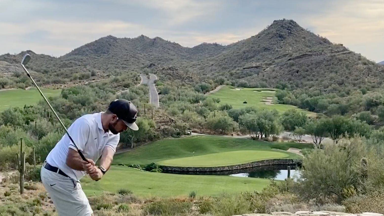 A man takes a swing at a tee that overlooks the rest of the golf course in a desert environment. 