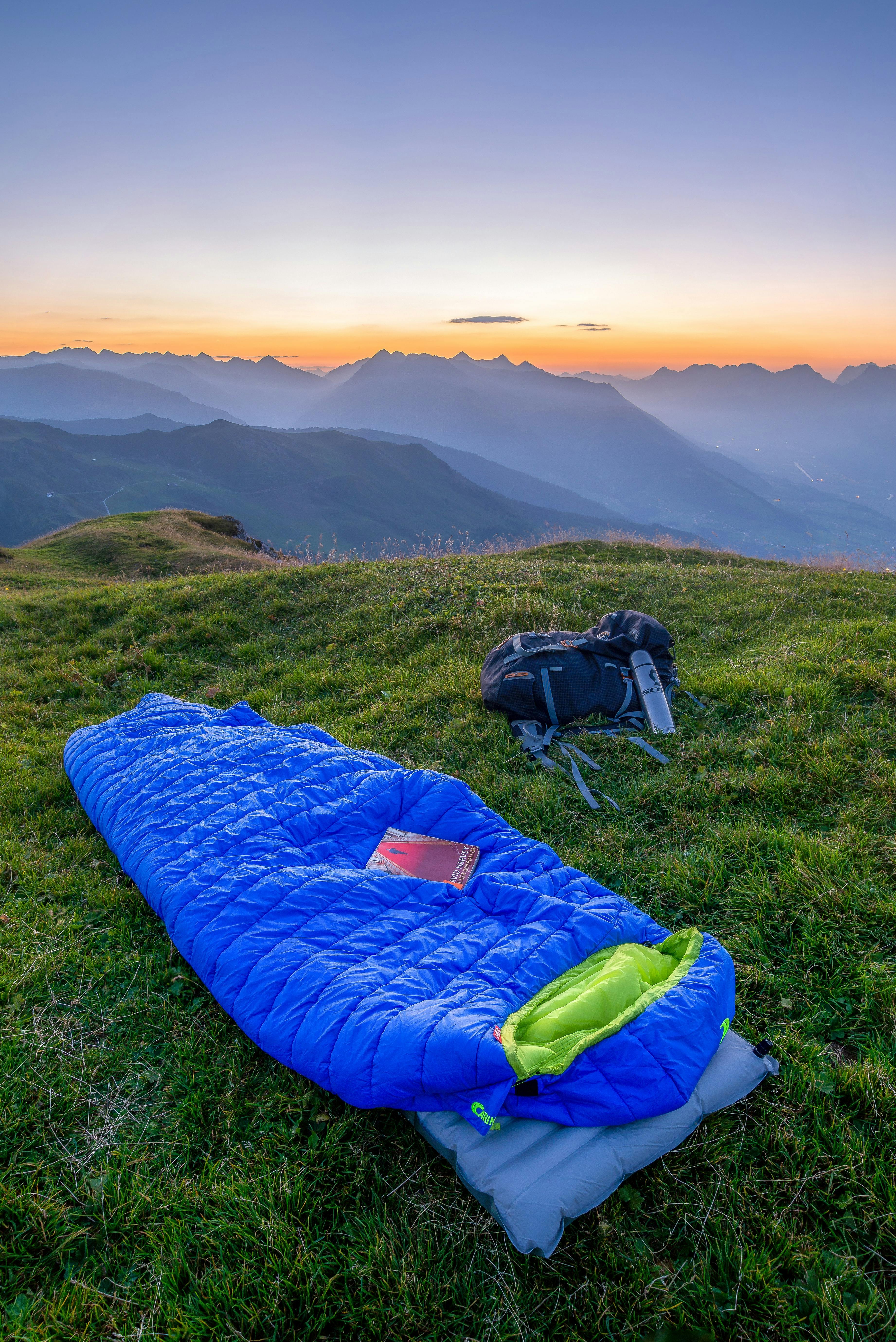 A blue sleeping bag is stretched out on a green hillside that overlooks a jagged ridge of mountains. 