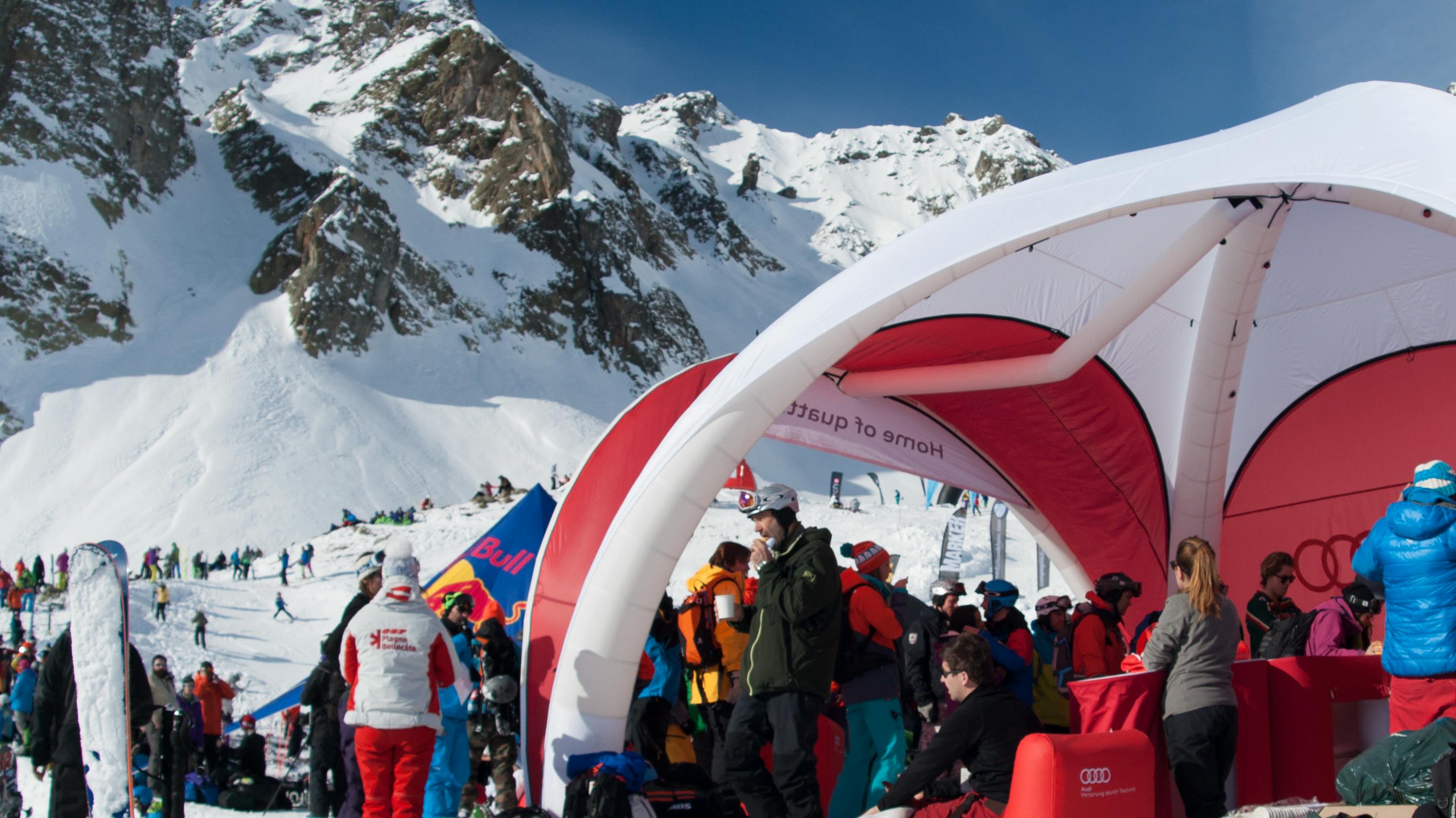 An Audi stand is set up on the snowy mountain for the Freeride World Tour. Skis stand upright in the snow and people mill about. 