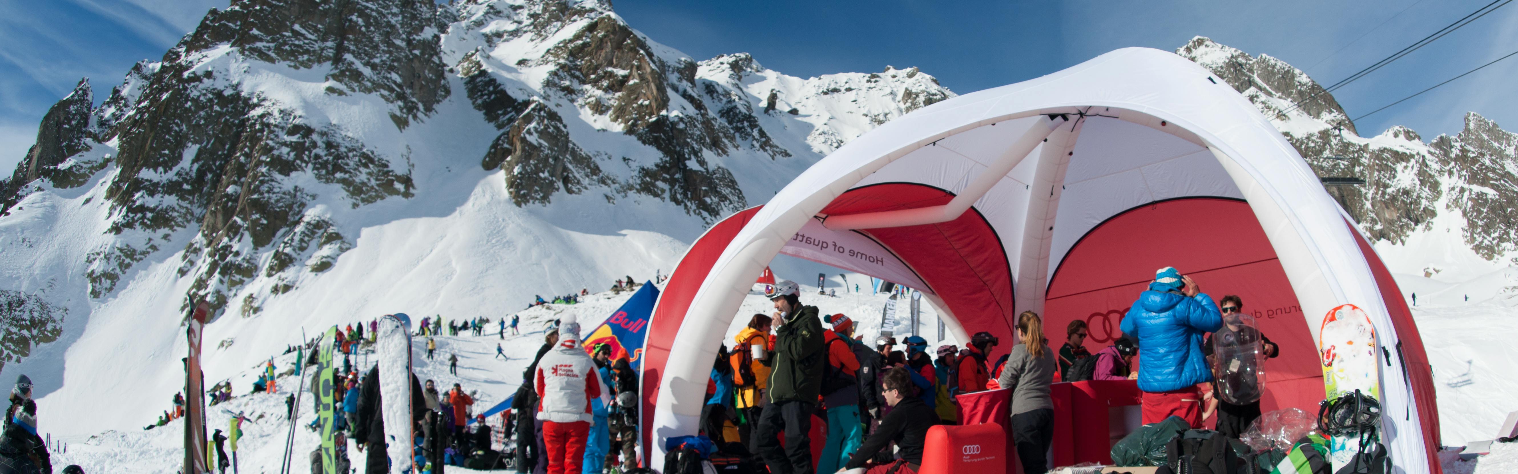 An Audi stand is set up on the snowy mountain for the Freeride World Tour. Skis stand upright in the snow and people mill about. 
