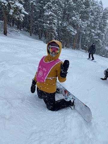 Snowboarder in yellow jacket and pink race bib waving at the camera while strapped into her board on a powder day.