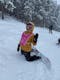 Female snowboarder in yellow jacket waves and kneels in the snow with her snowboard attacked on a powder day.