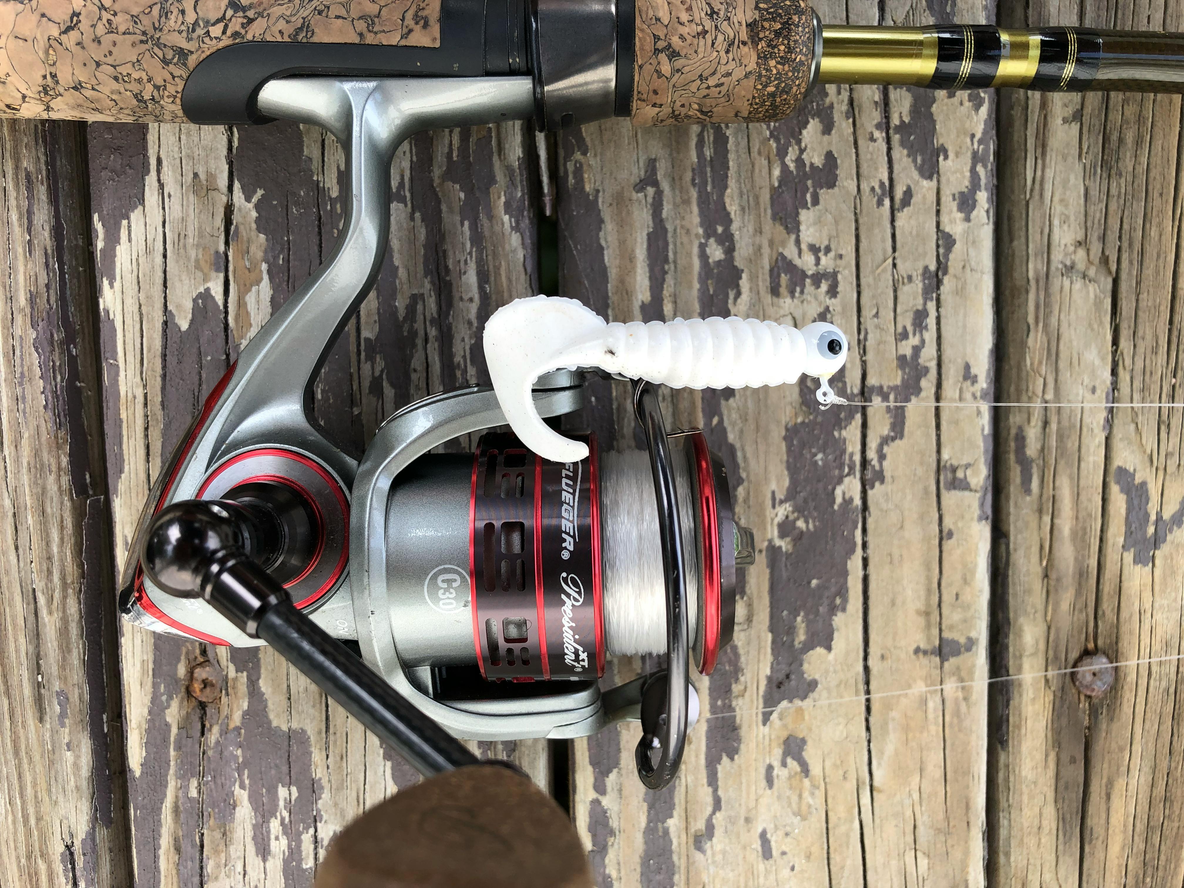 A spinning reel and rod lay on a wooden table with a white twister tail jig strung on the line. 