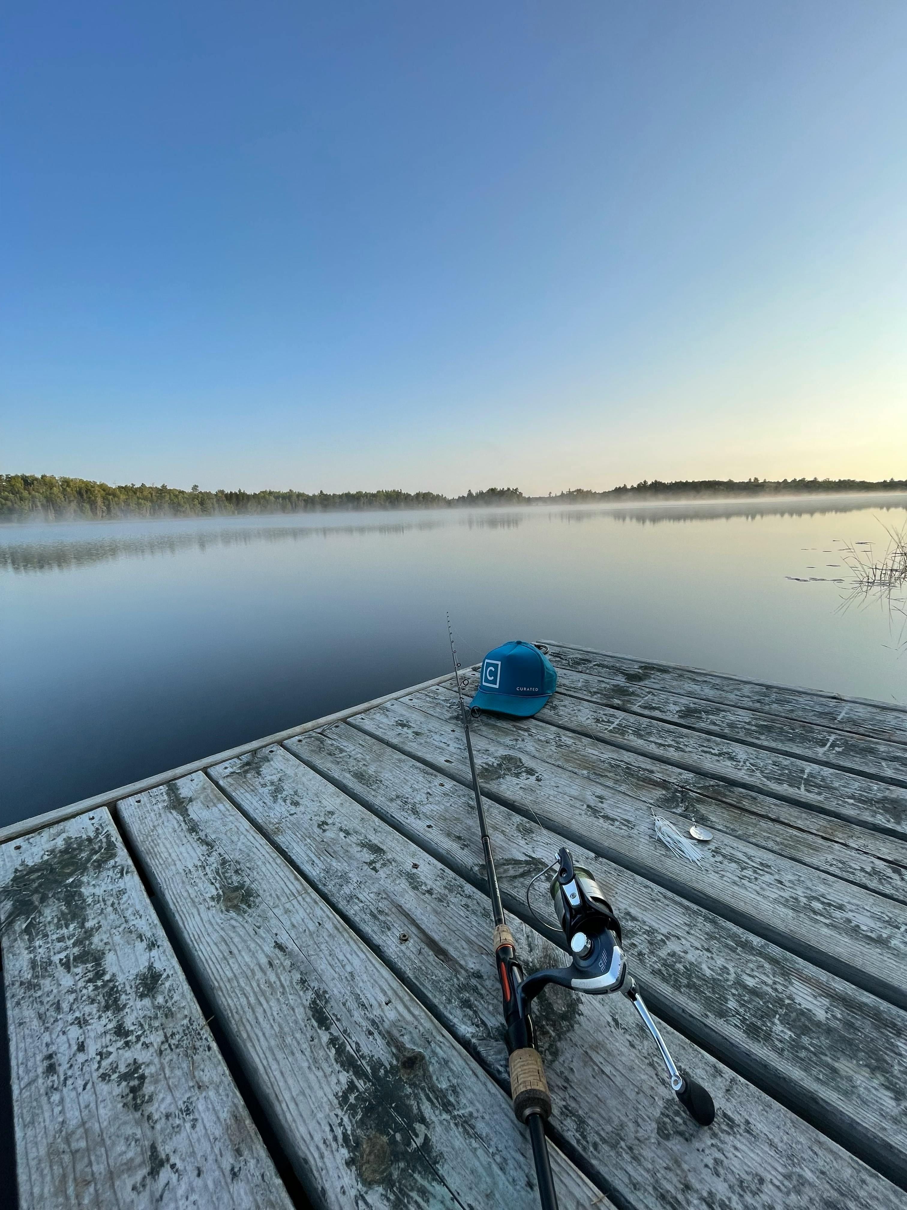 A view from a dock on a lake with a fishing rod and blue Curated hat on it. 