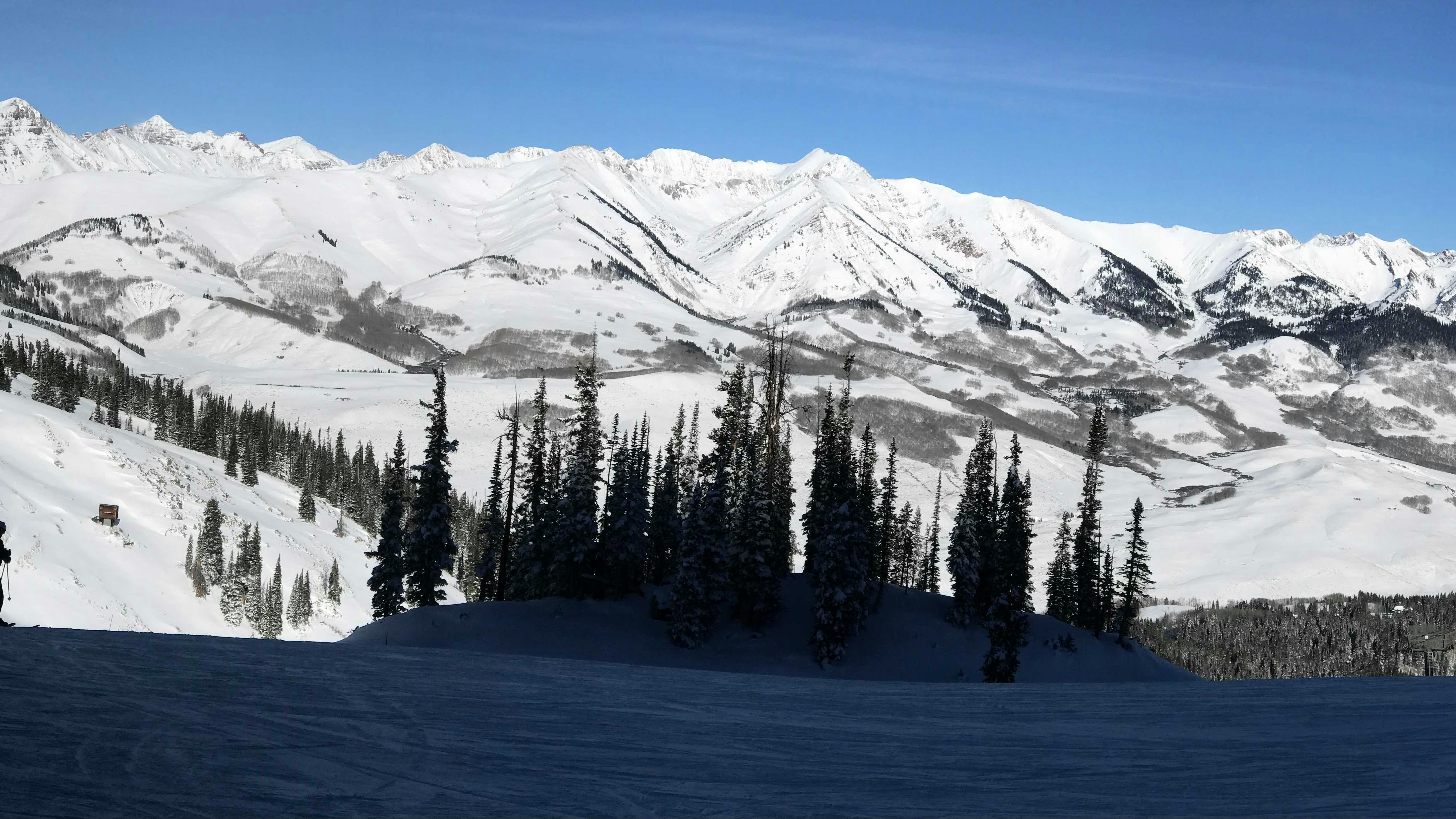 A panorama of a ski slope with someone skiing across in the shade of the mountain.
