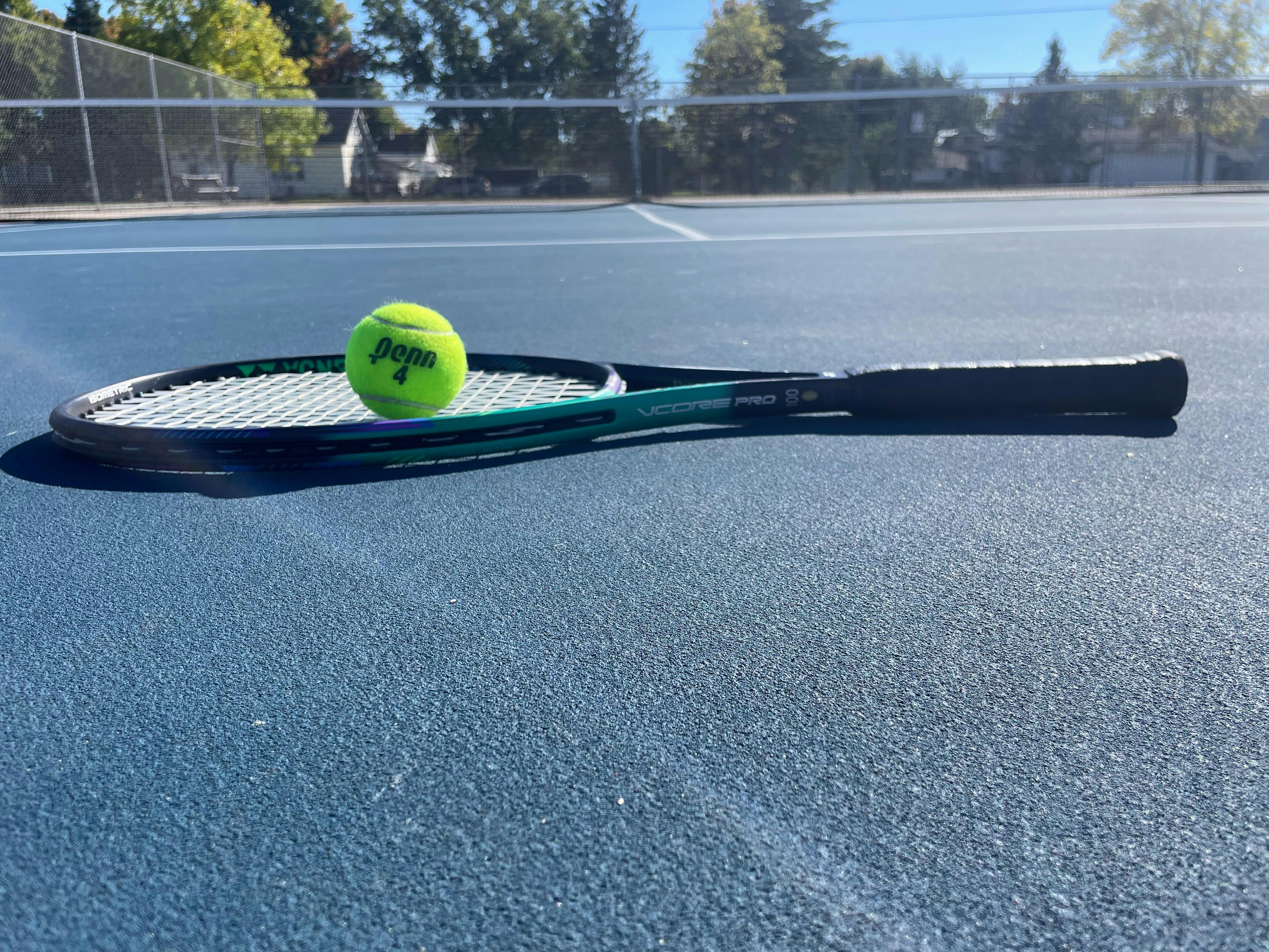 A Yonex VCore Pro 100 Racquet · Unstrung with a tennis ball on it laying on the court. 