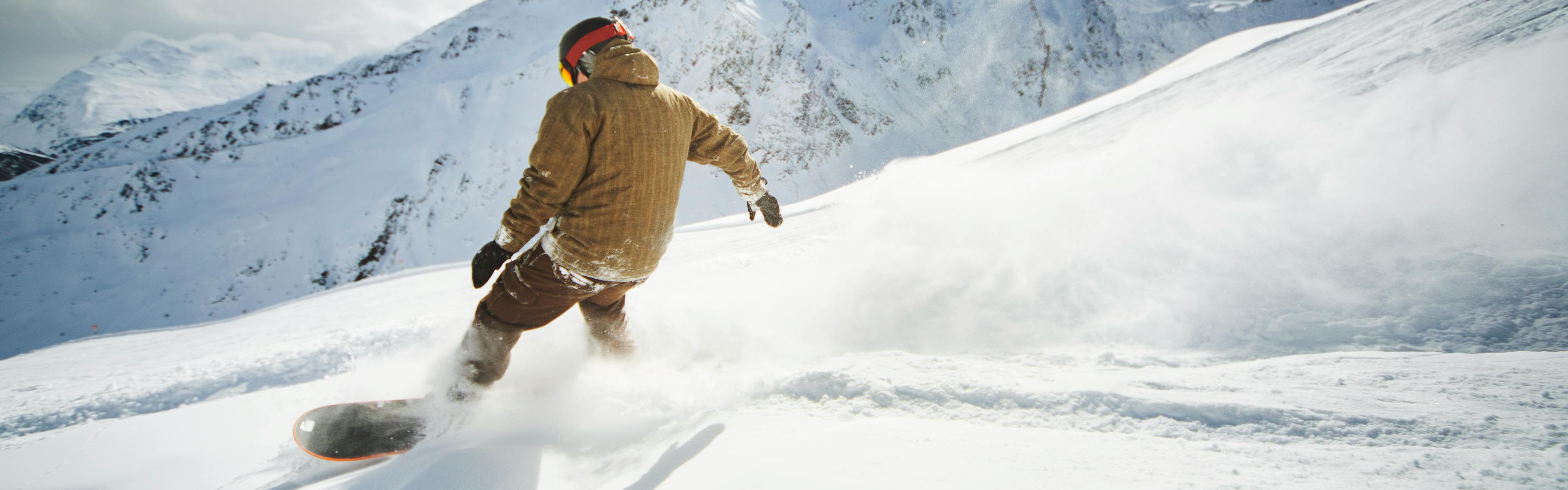 A snowboarder in a beige jacket makes their way down the mountain with a cloud of powder behind them