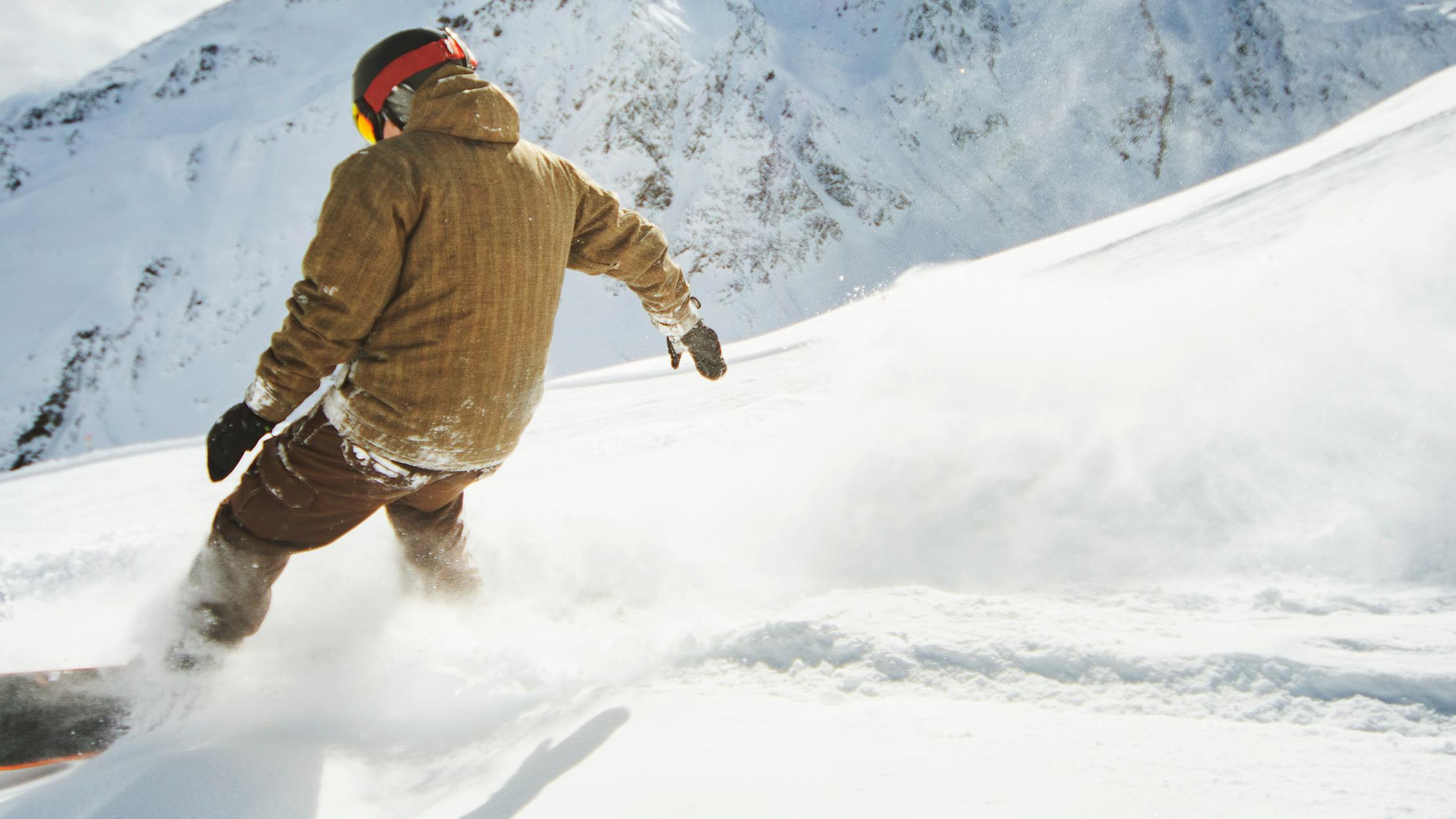 A snowboarder in a beige jacket makes their way down the mountain with a cloud of powder behind them