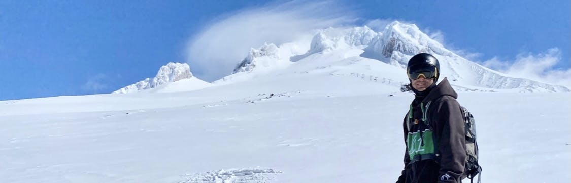 A skier stands with a helmet and goggles on. There is a snowy mountain and blue skies in the background. 