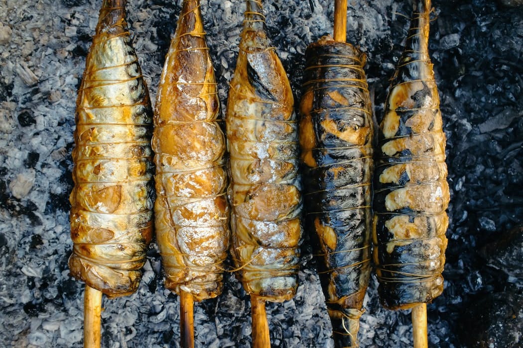 Image of five fish bodies skewered and tied in twine over the hot coals of a fire.