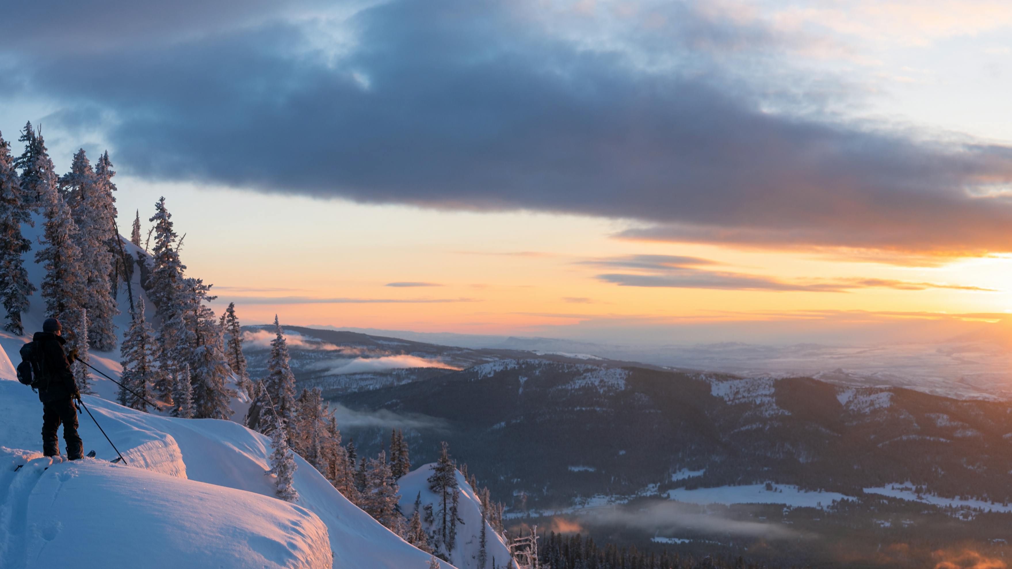 Two people on skis stand on a snowy hill and look out over the sunrise.