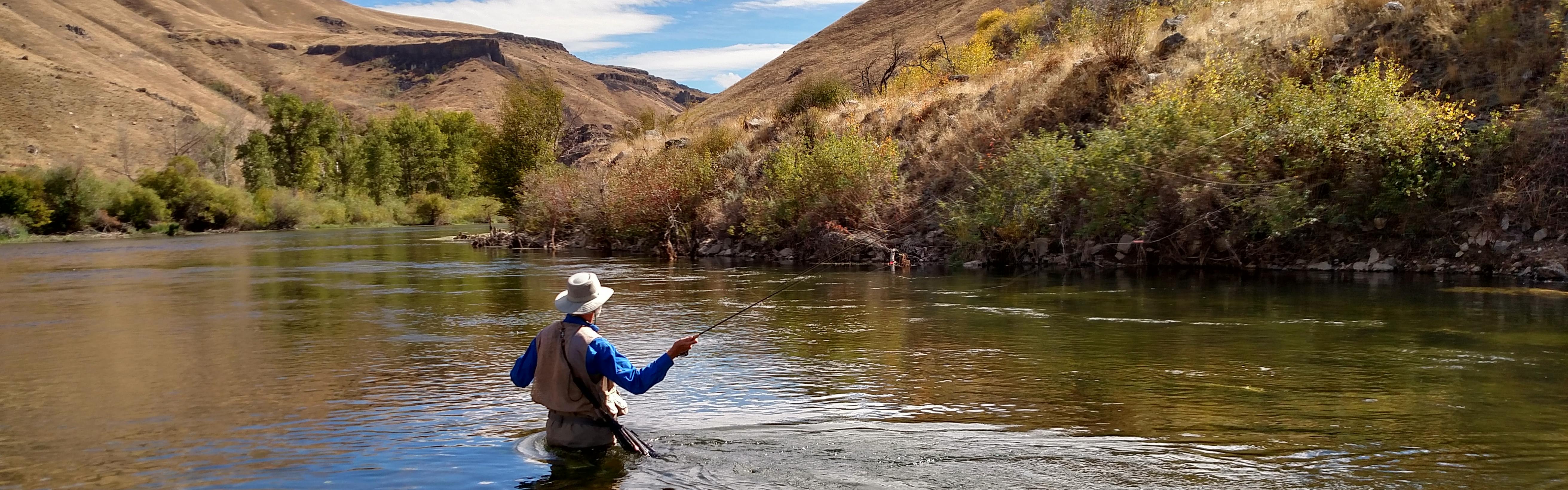 A fly fisher stands in a river with a vest and hat on.