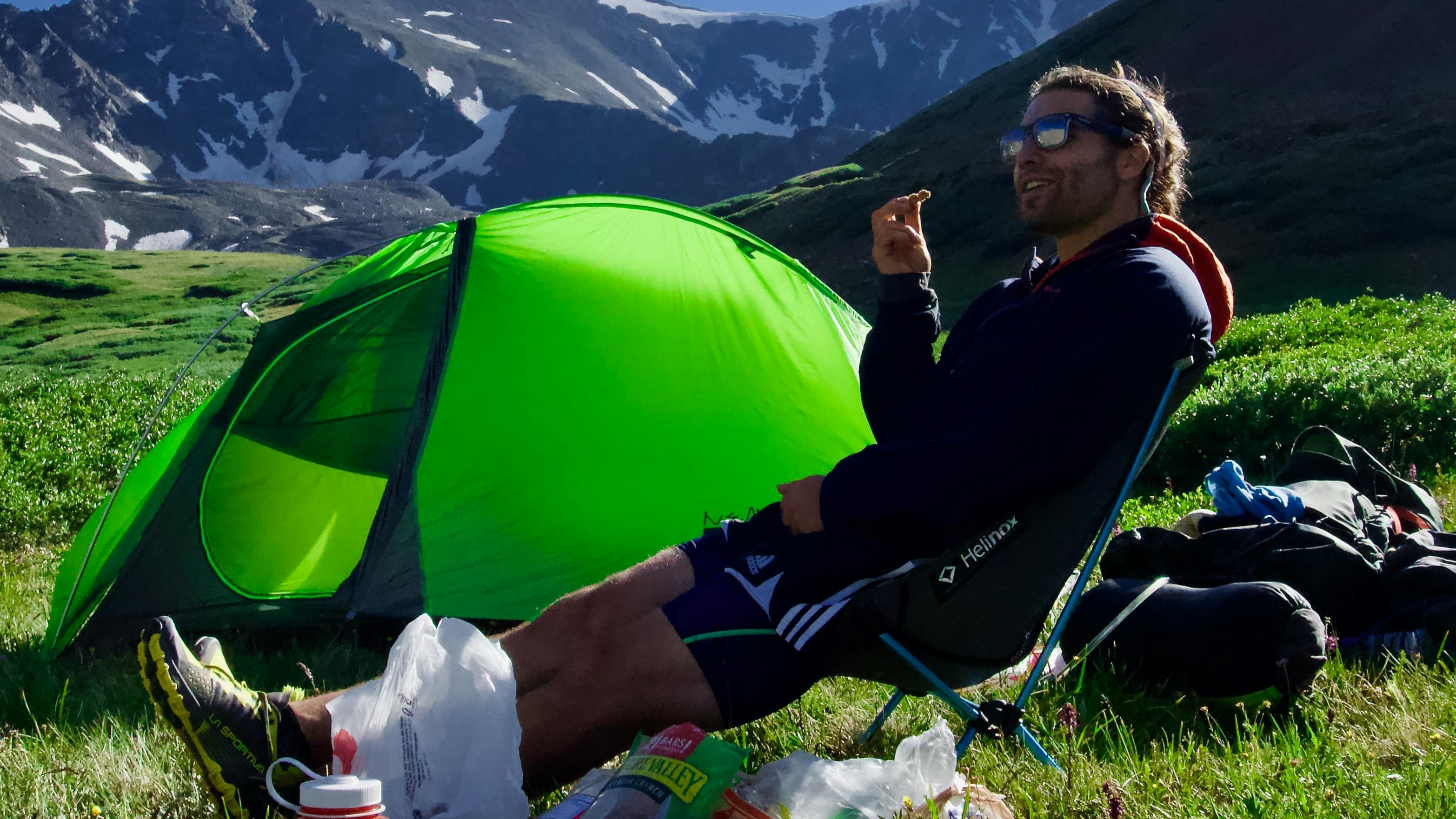 A man eating some food near his tent. There is a snowy mountain in the background. 