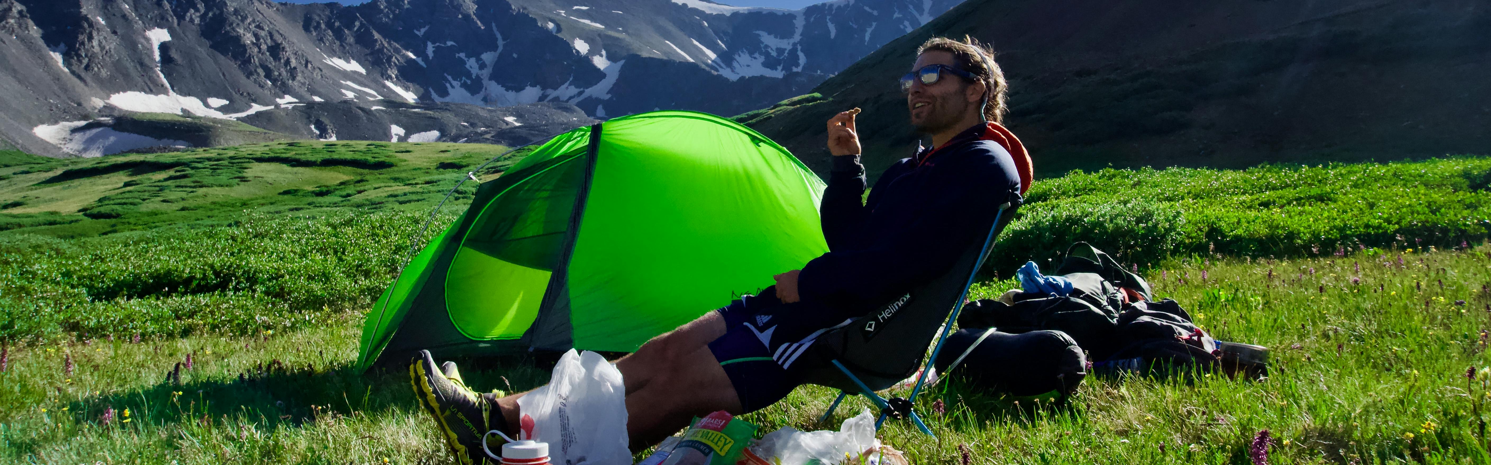 A man eating some food near his tent. There is a snowy mountain in the background. 