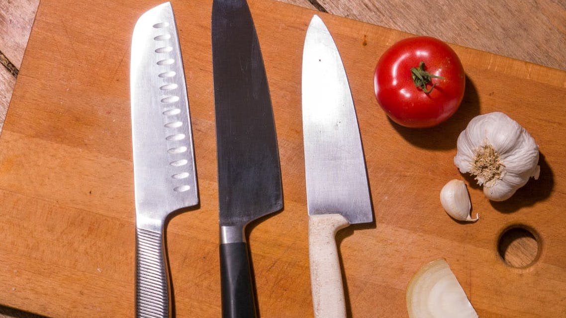 Three knives in a row on a cutting board. There is a tomato and some garlic on the cutting board also. 