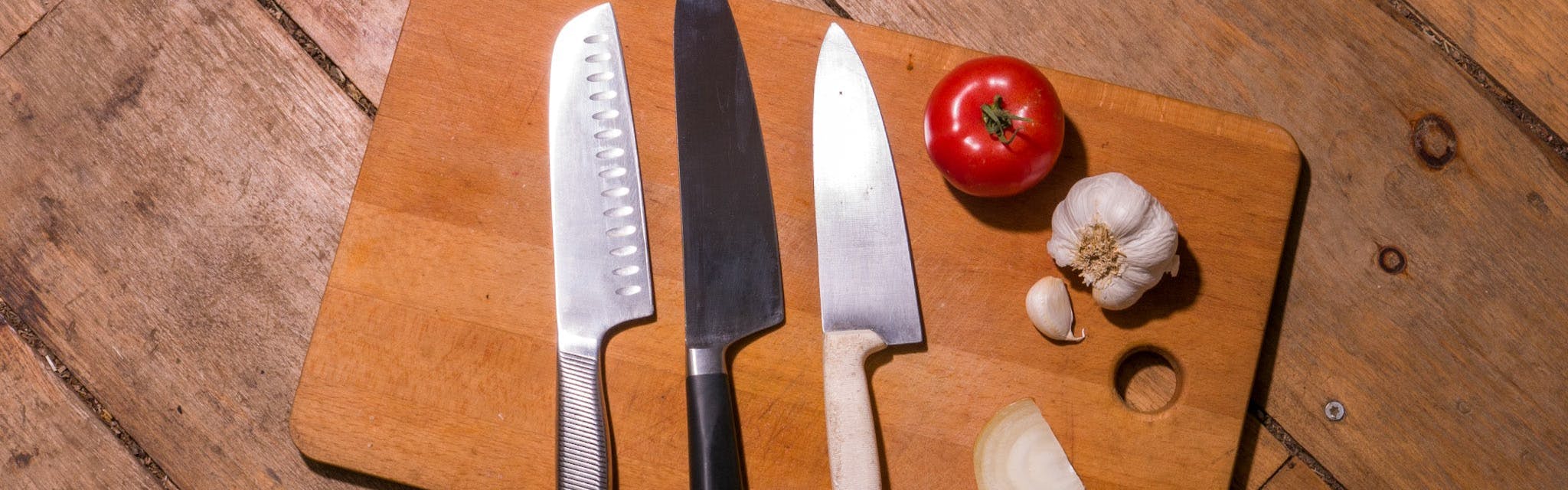 Three knives in a row on a cutting board. There is a tomato and some garlic on the cutting board also. 