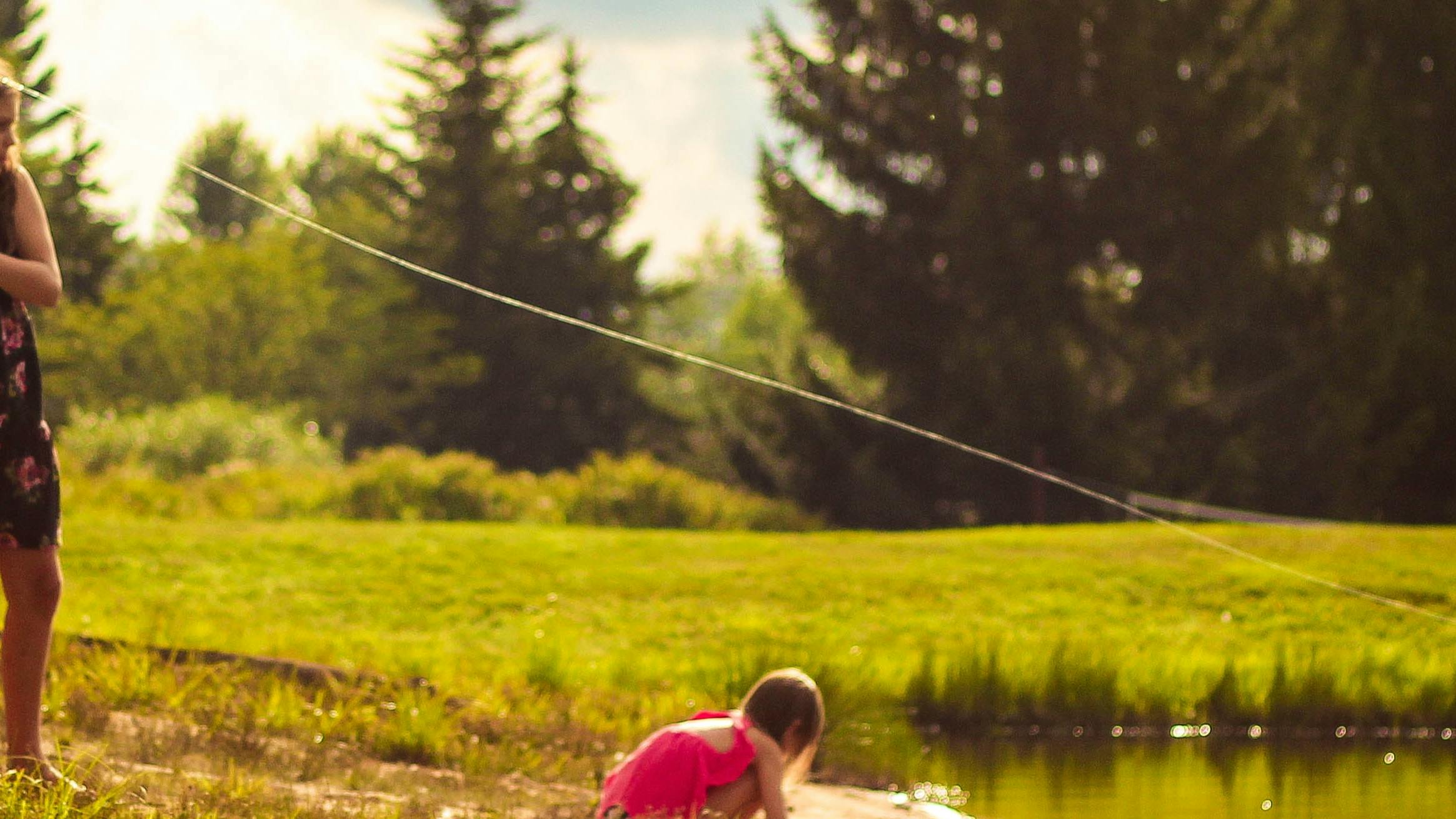 A girl in a dress stands barefoot on the grassy banks of a pond and pulls back a fishing rod. Another younger girl crouches and plays in the mud.