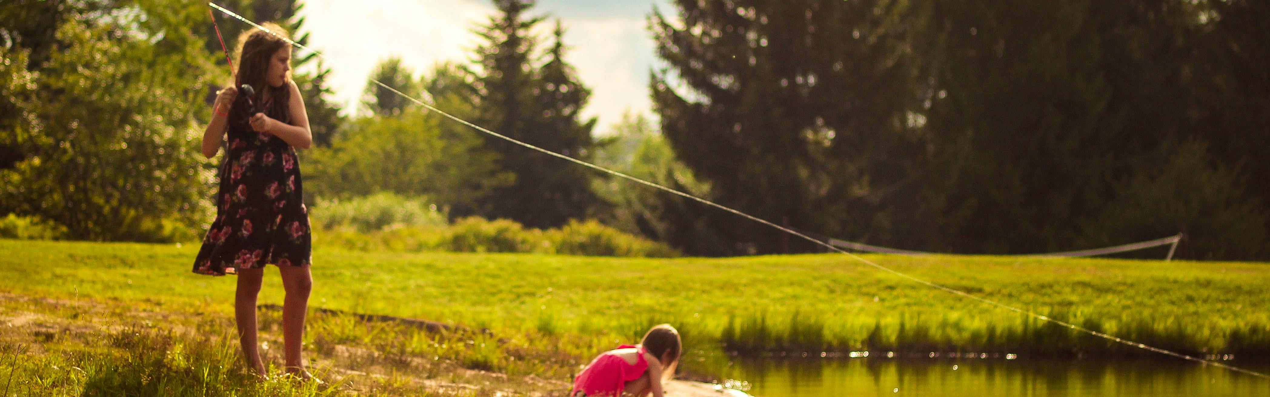 A girl in a dress stands barefoot on the grassy banks of a pond and pulls back a fishing rod. Another younger girl crouches and plays in the mud.