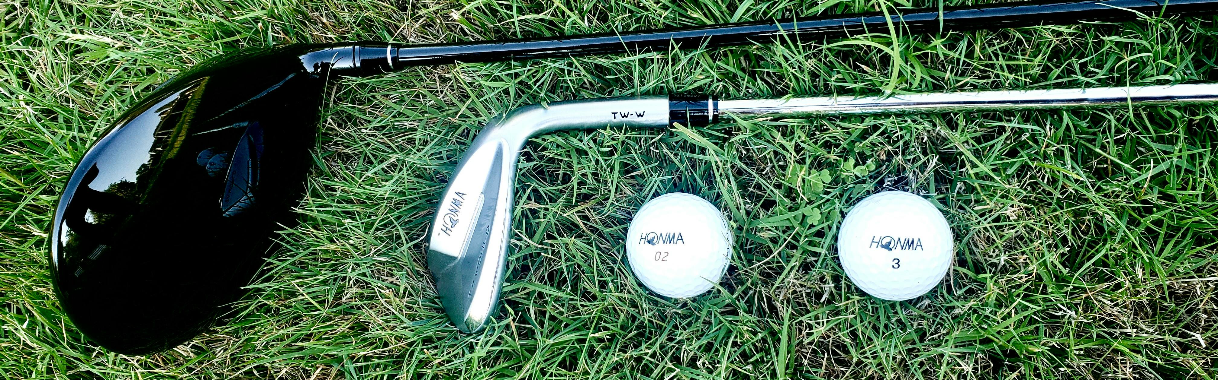 A black golf club and a silver golf club lie on the grass next to two Honma golf balls