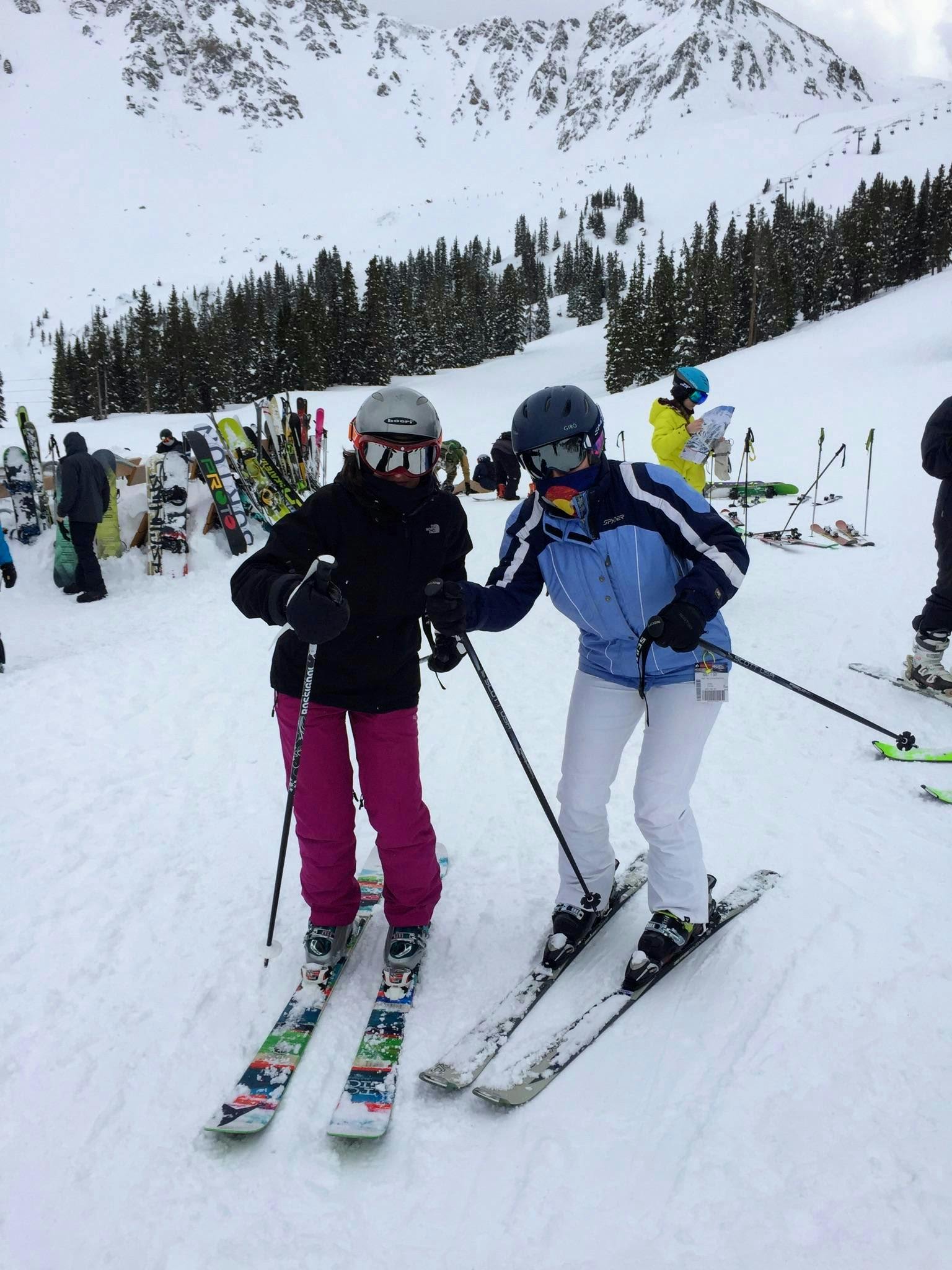 Two skiers standing on a ski run. There is a snowy mountain in the background.