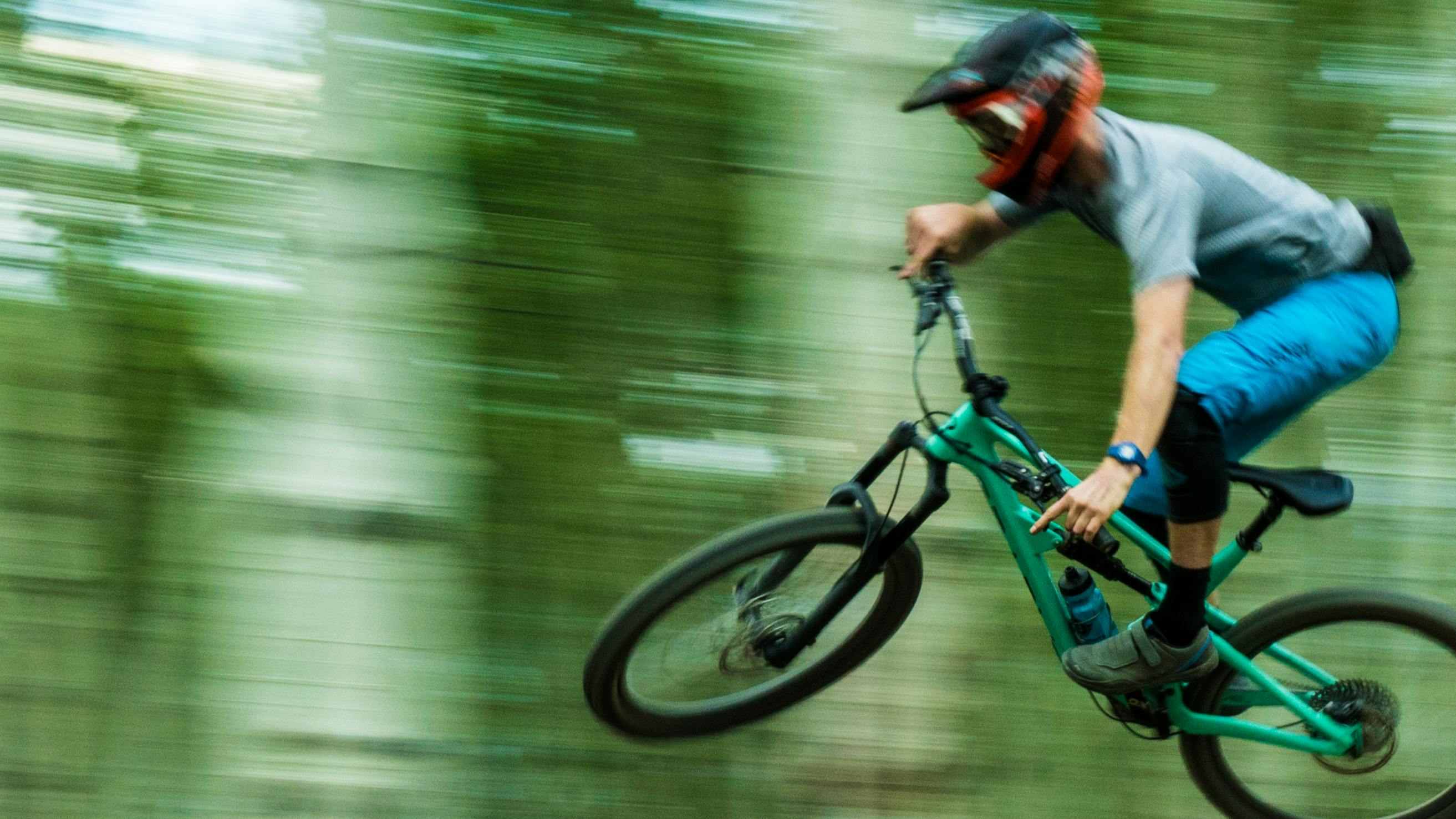 Man on mountain biking flying through the air, the green forest a blur behind him.