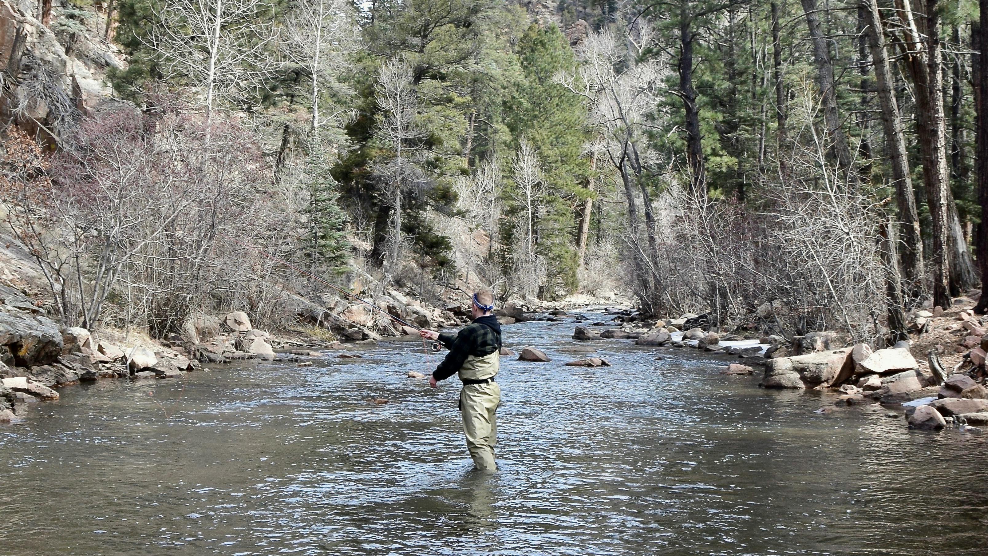 A man flyfishes in the middle of the river while wearing waders. The banks are sloped and rocky. 