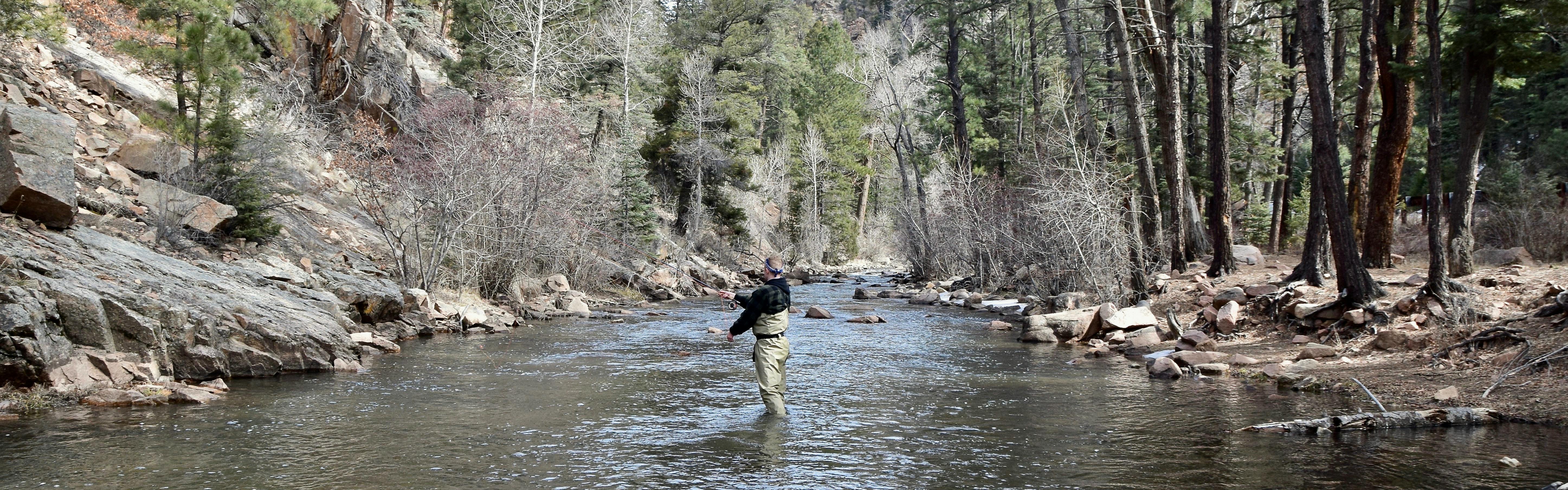 A man flyfishes in the middle of the river while wearing waders. The banks are sloped and rocky. 