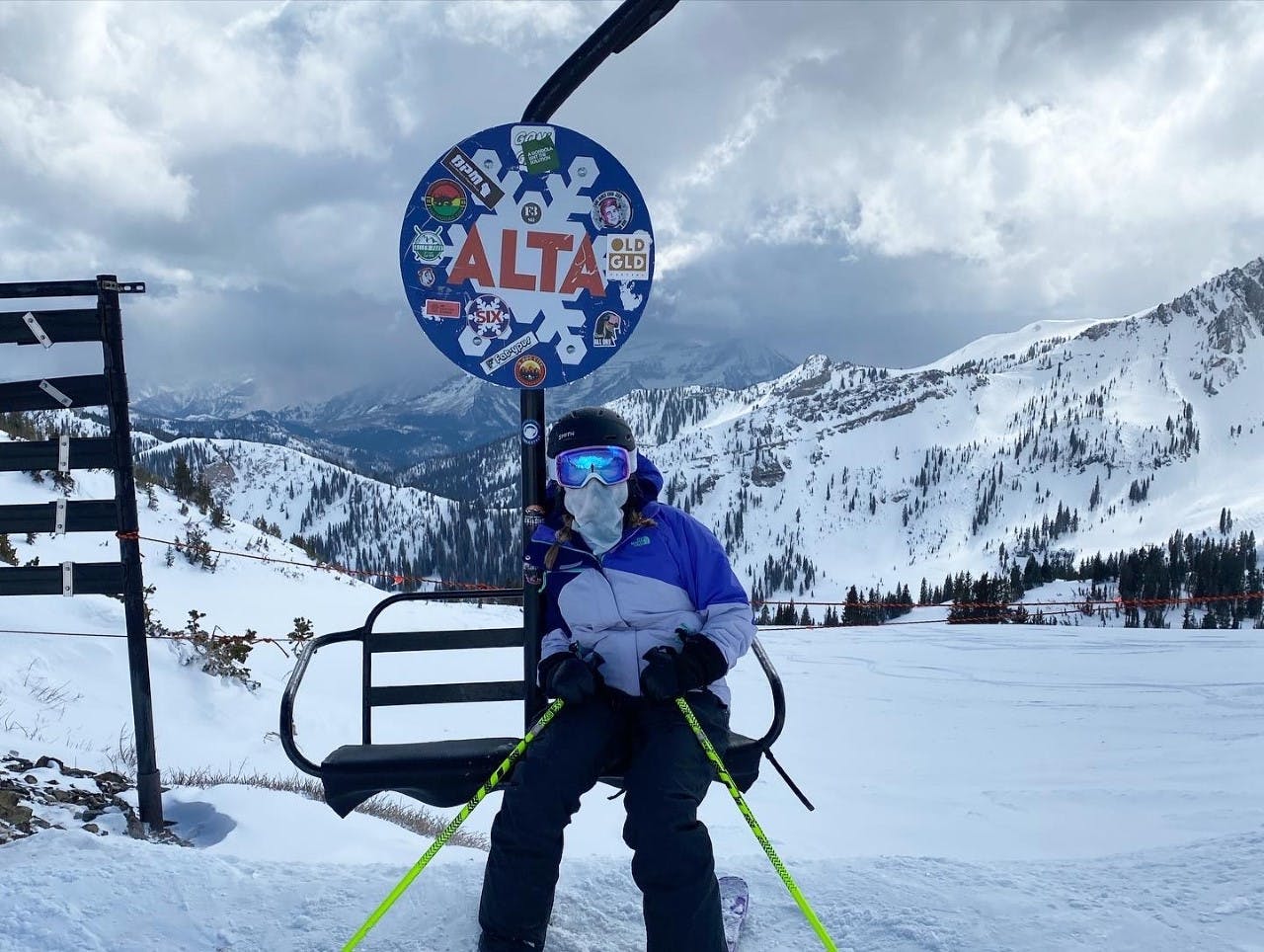 A skier with poles, a helmet, and goggles sits under a sign on a ski run that says "Alta".