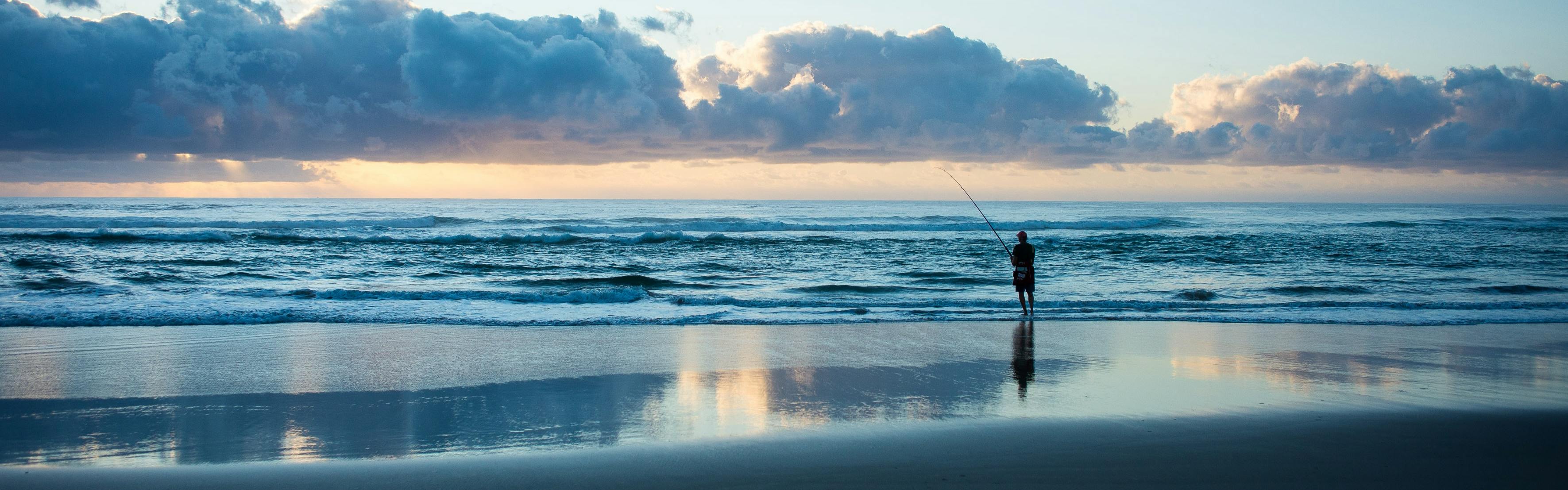 A fisherman standing on the shore of the ocean with a fishing rod. The sun is starting to come up. 