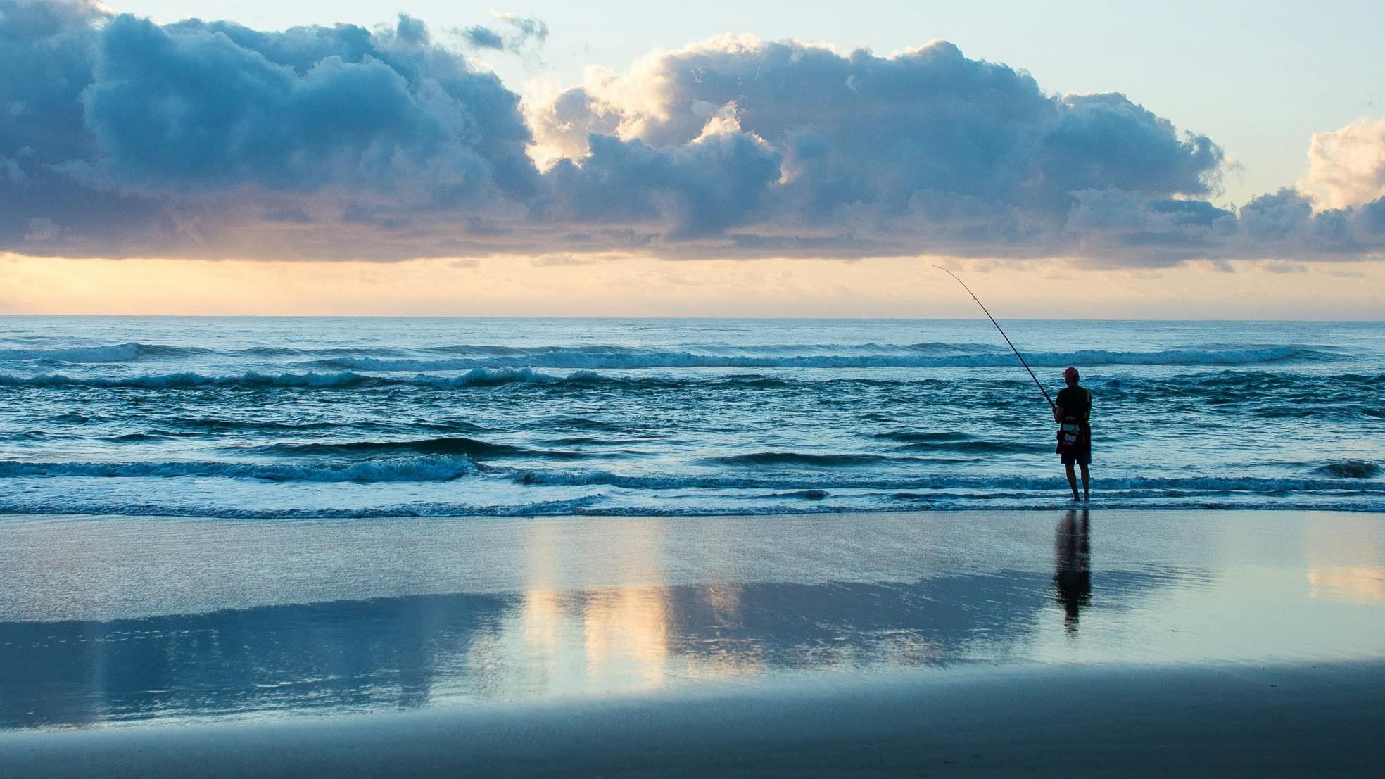 A fisherman standing on the shore of the ocean with a fishing rod. The sun is starting to come up. 
