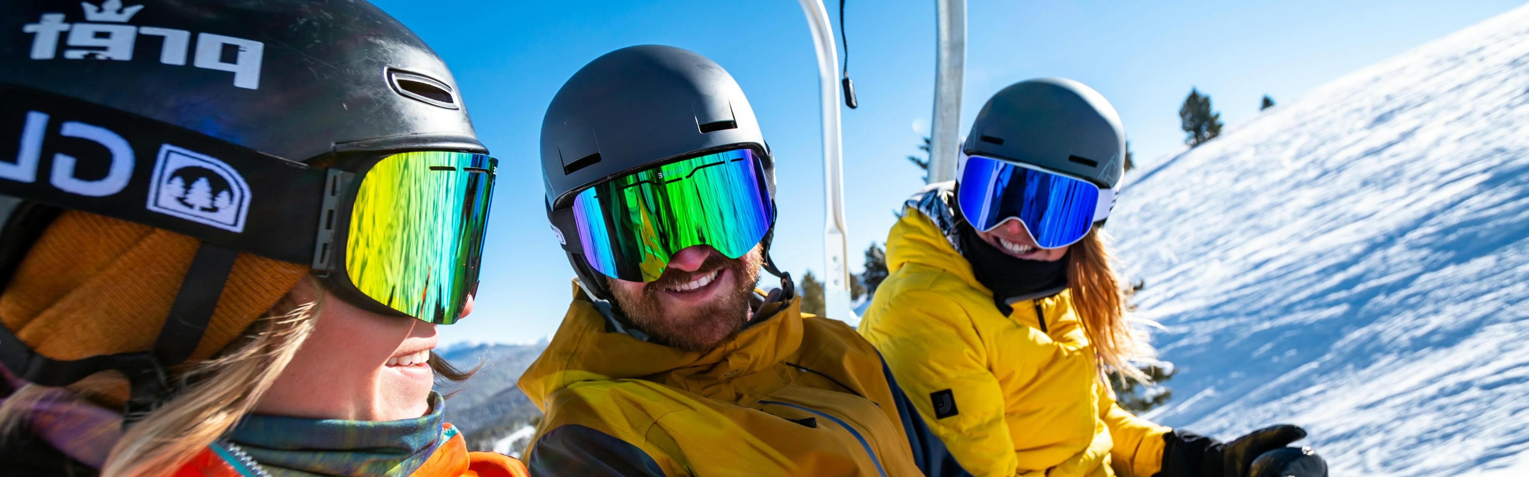 Three people on a chair lift wear ski goggles and helmets and smile at each other.