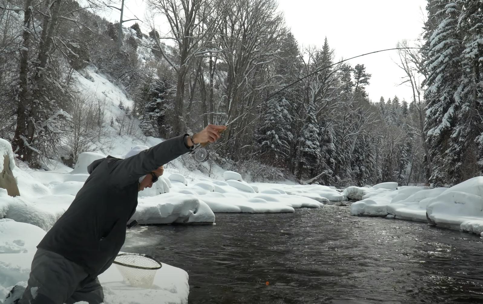 A fisherman on a snowy riverbank with a fishing rod. 