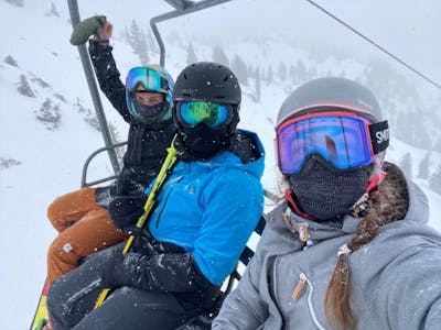 Three people sit on a chairlift at a ski resort. It is snowing. 
