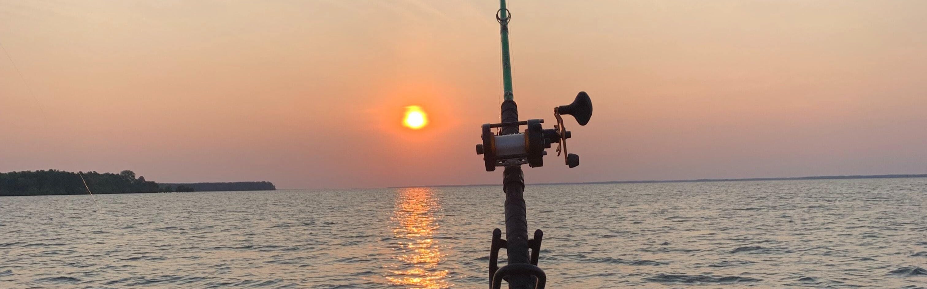 Someone has a baitcasting rod set up off the back of a boat at sunset. 