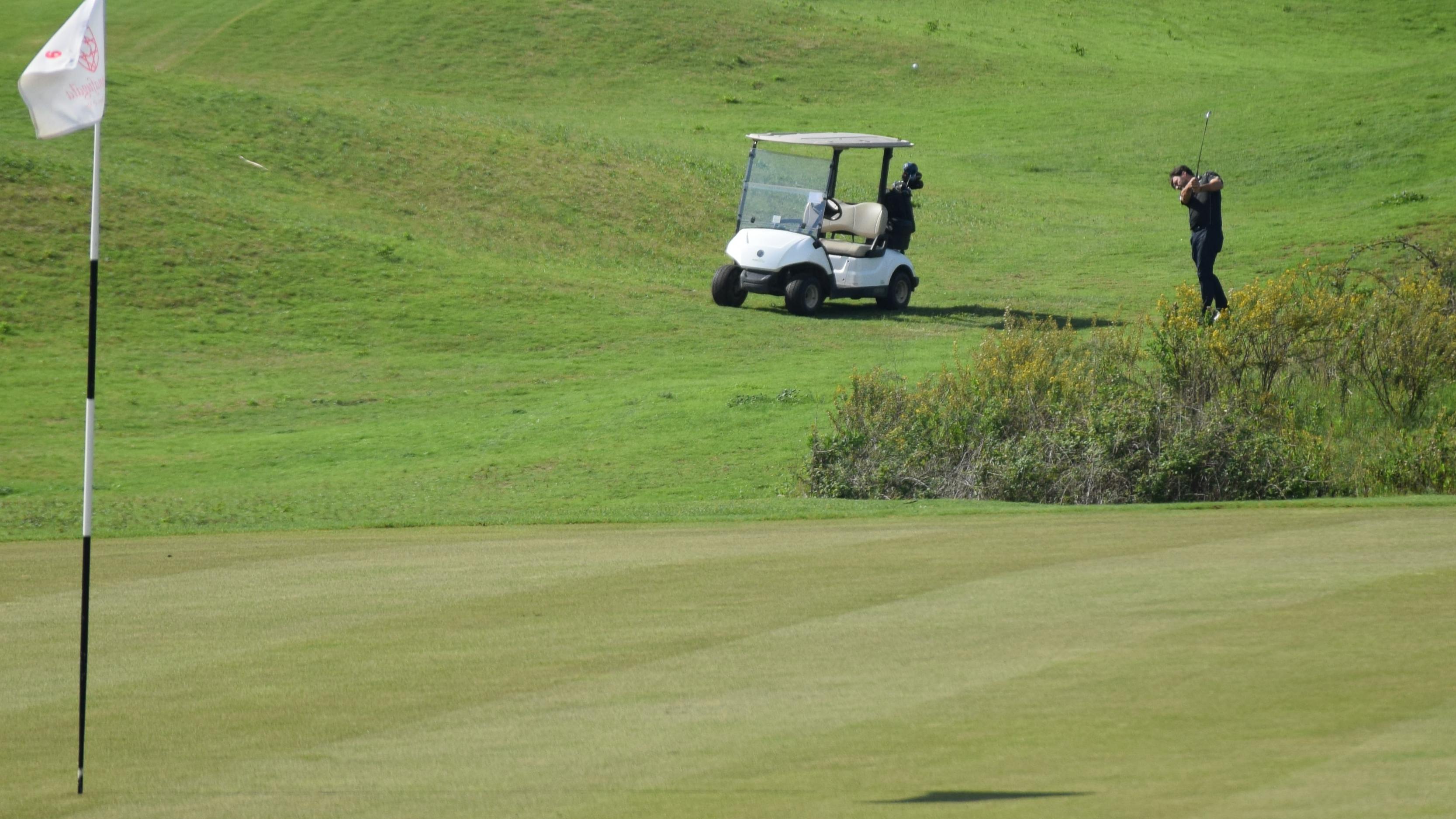 A golfer takes a swing towards the pin. His golf cart is behind him.