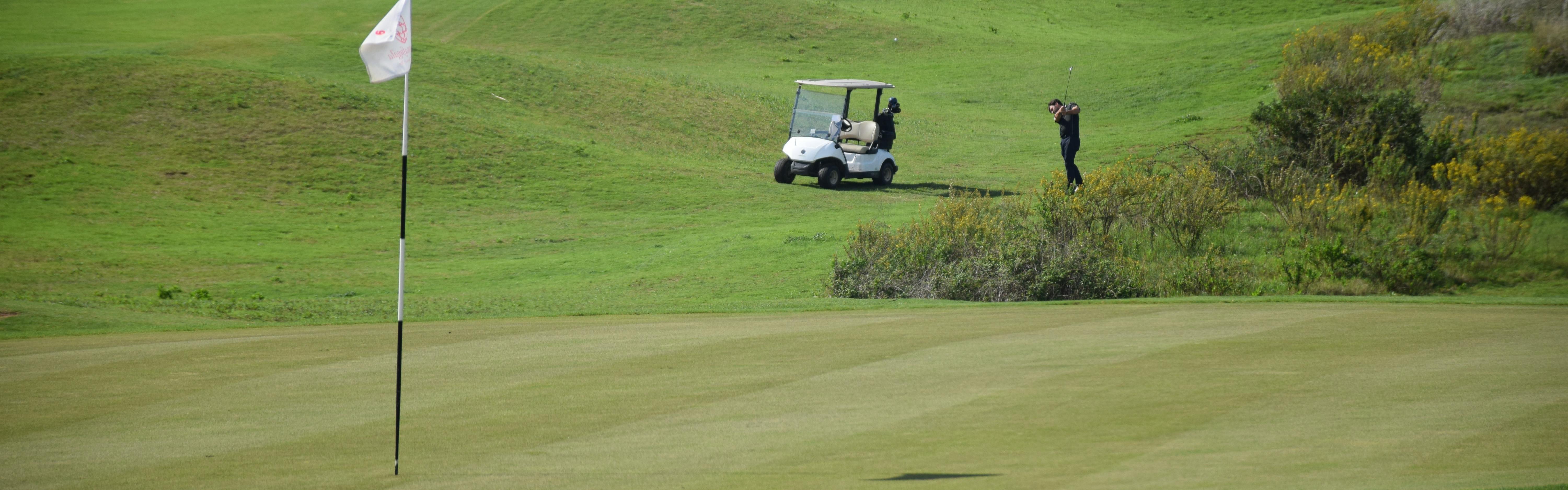 A golfer takes a swing towards the pin. His golf cart is behind him.
