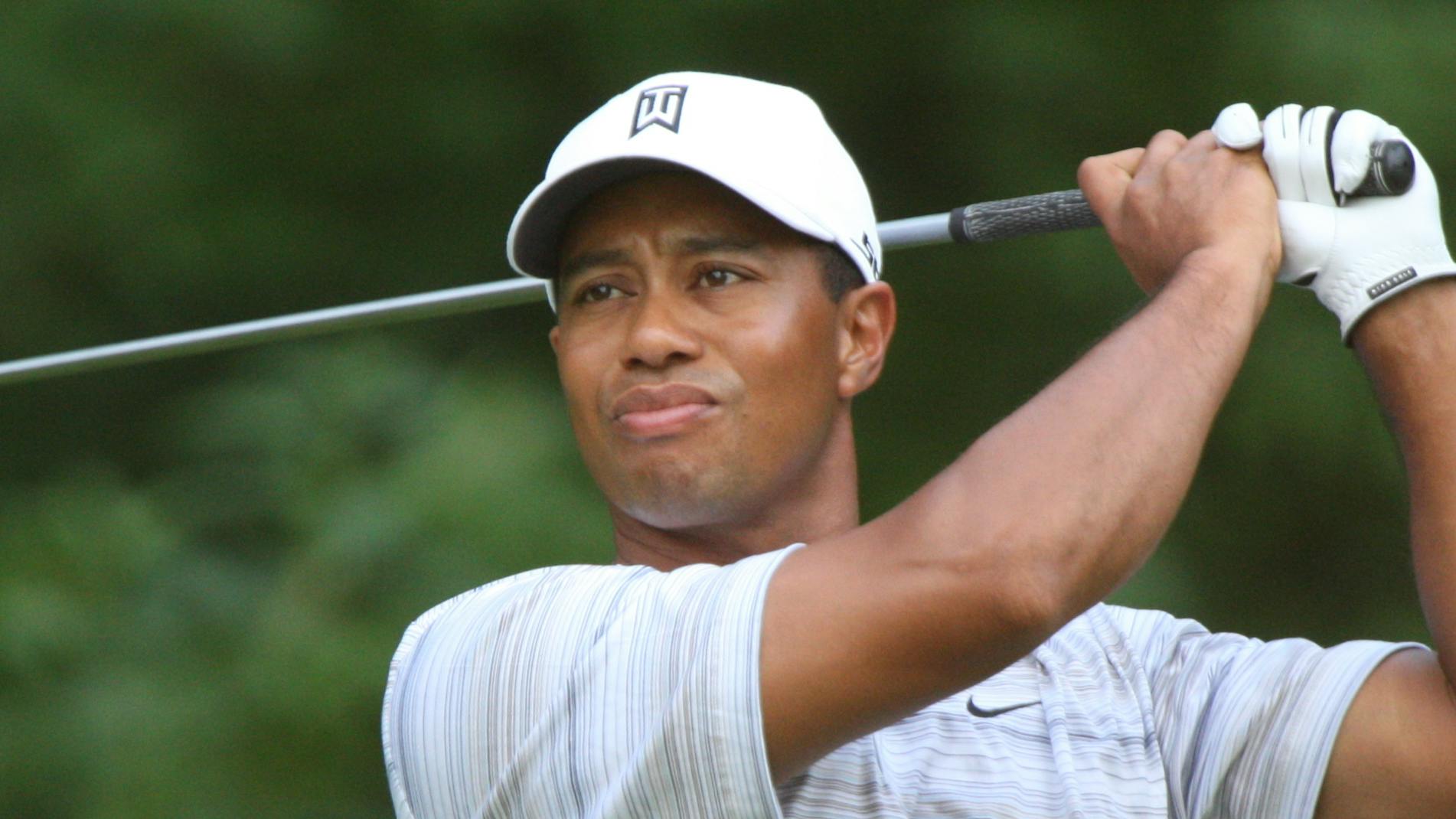 Tiger Woods swings back a driver while wearing a white hat. 