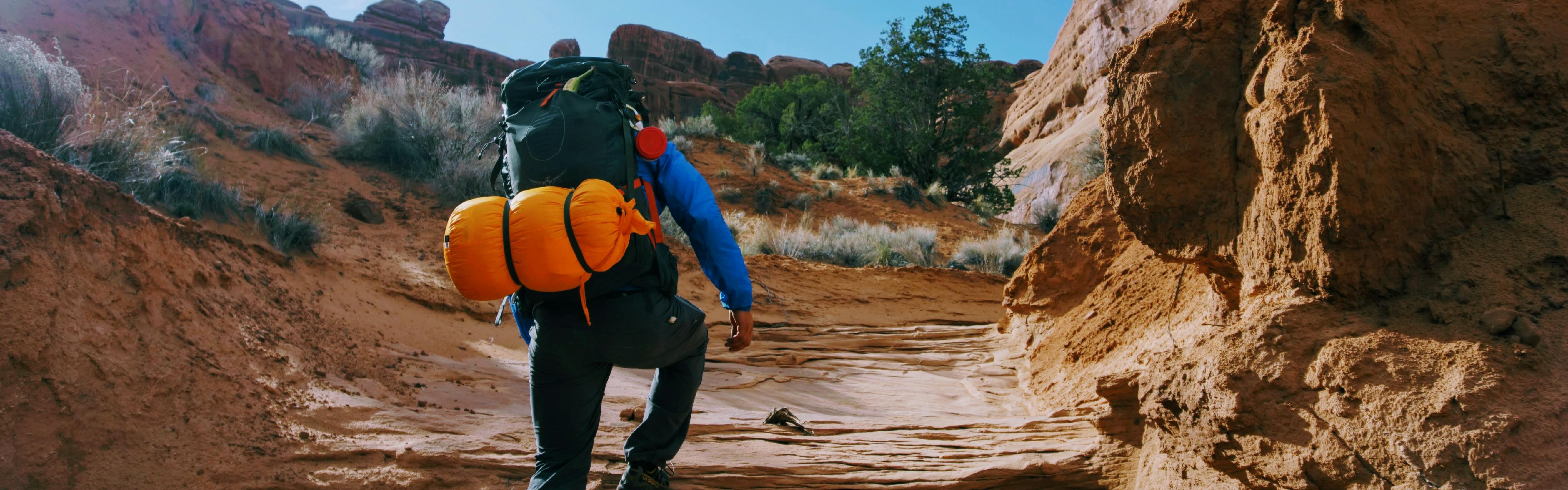 A backpacker walking up a rocky trail with his sleeping bag attached to the outside of his bag. 