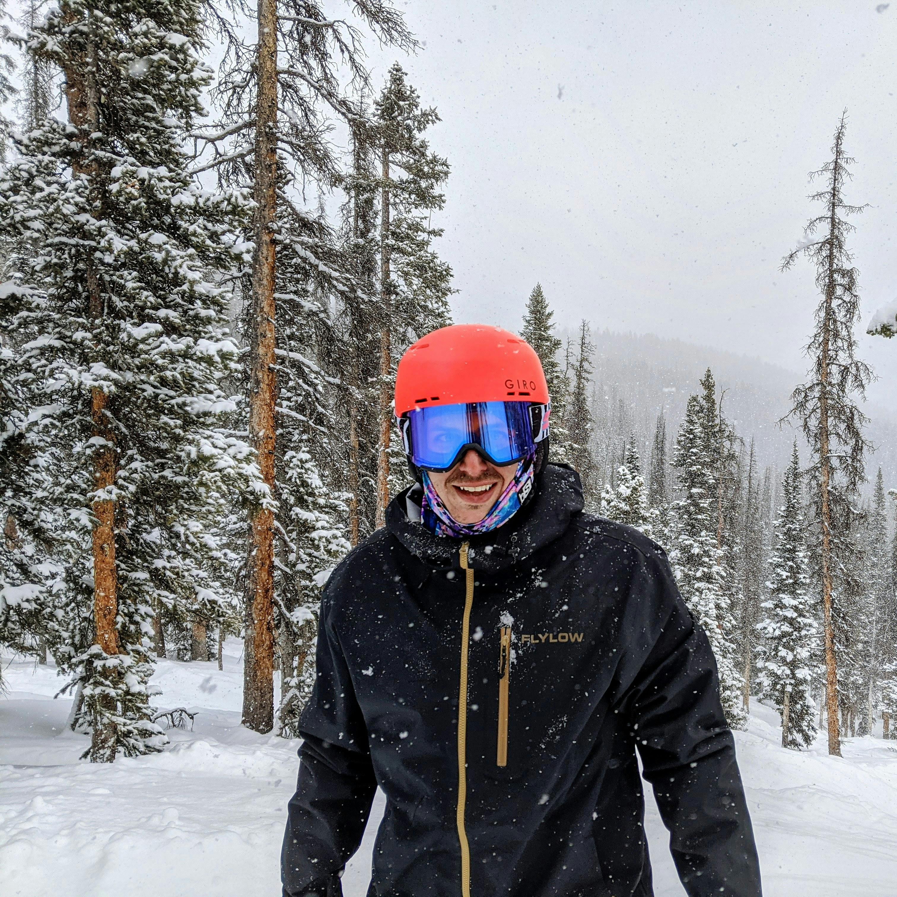 A skier wearing the Giro Emerge helmet smiling at the camera. There is snow falling and snowy trees behind him. 