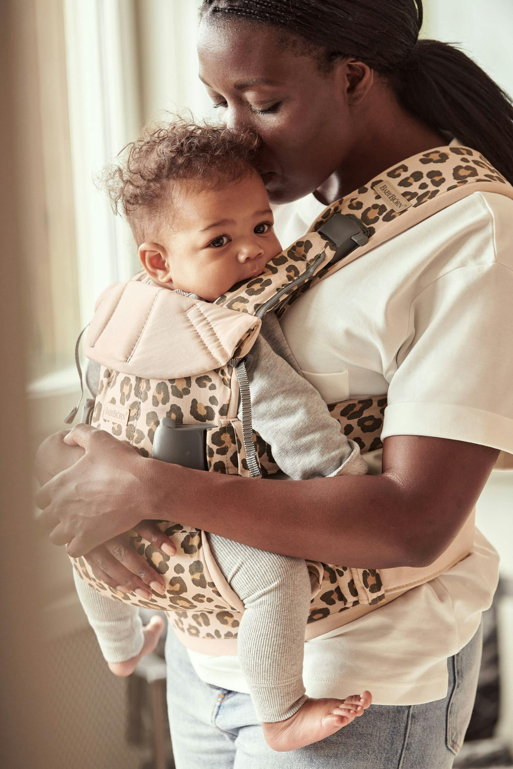 A woman holds a baby in the Baby Carrier One on her front and tips her head down to kiss the baby's head. 