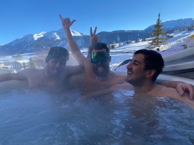 Three men in a hot tub. One is wearing ski goggles. There is a snowy mountain in the background. 