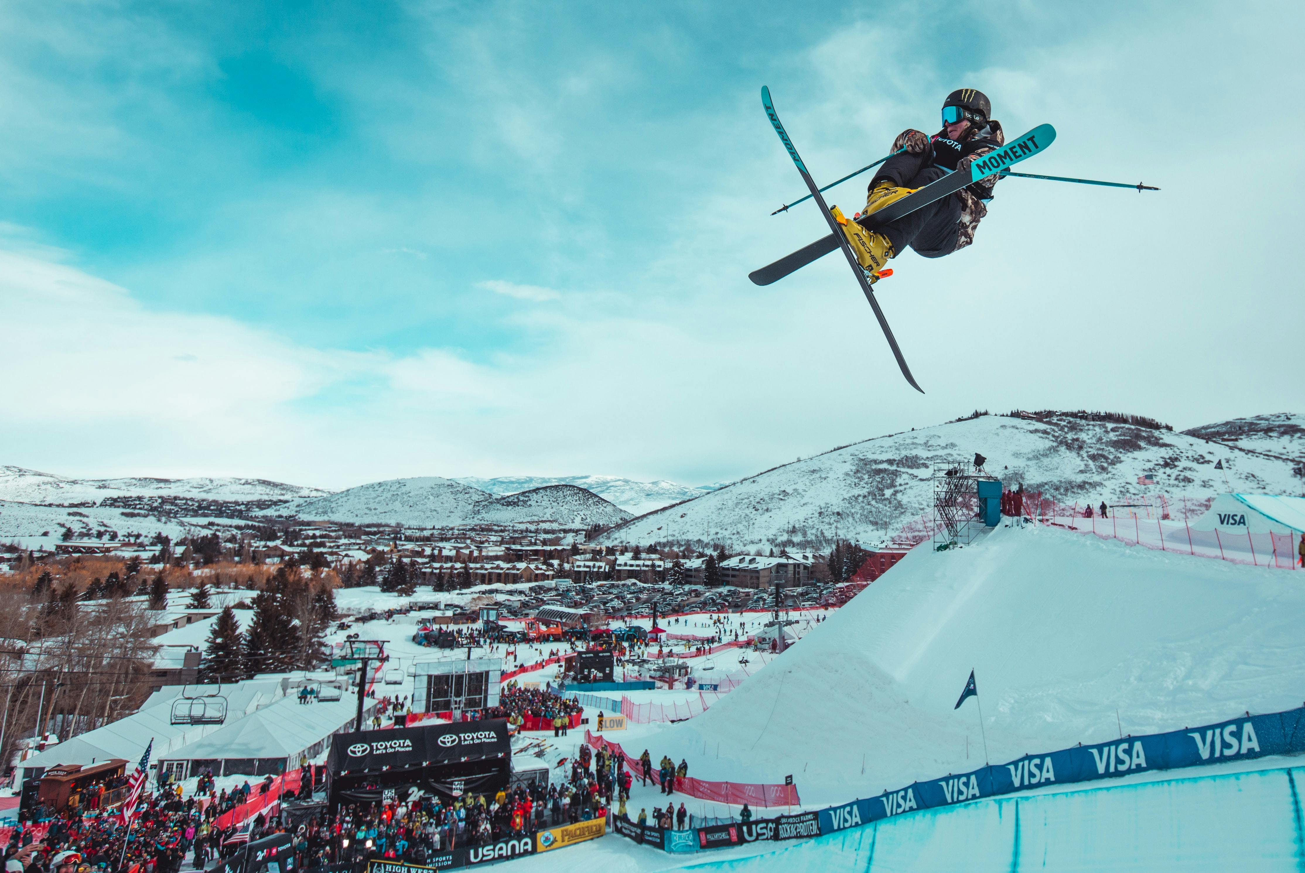 A skier doing a jump and grabbing his skis. There is a crowd in the background watching him. 