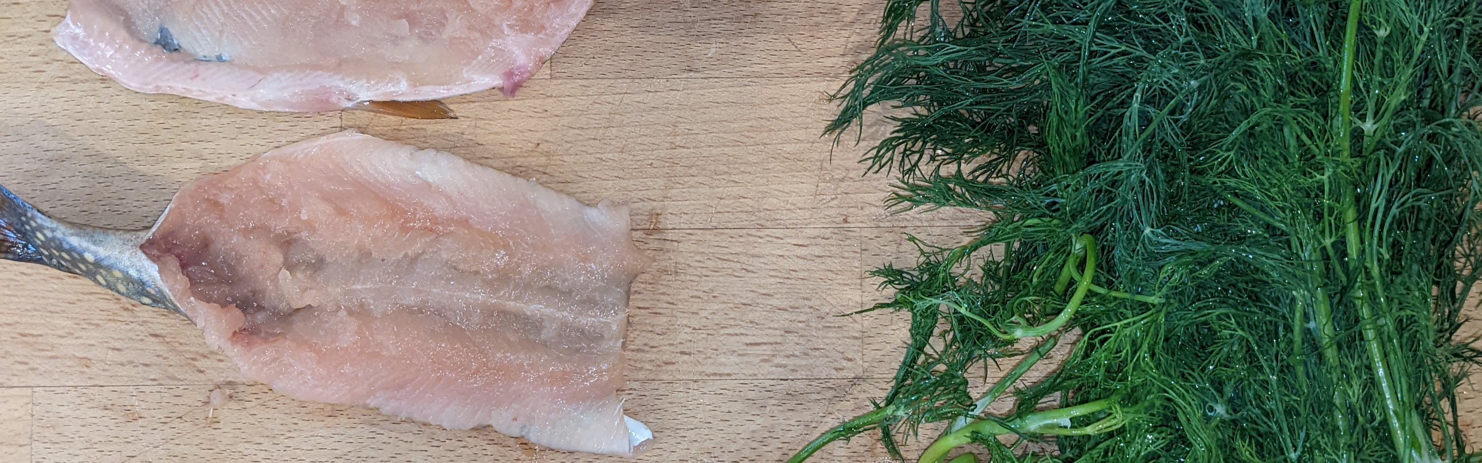 A fish fillet lies on a cutting board with some herbs.