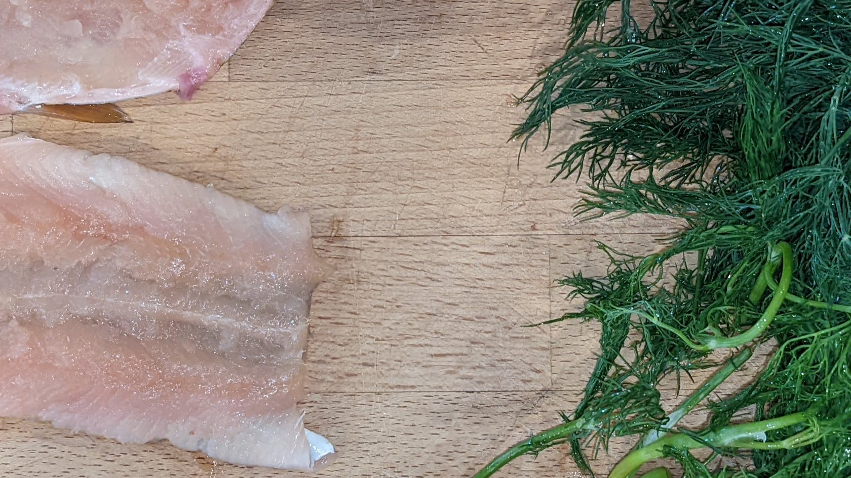 A fish fillet lies on a cutting board with some herbs.