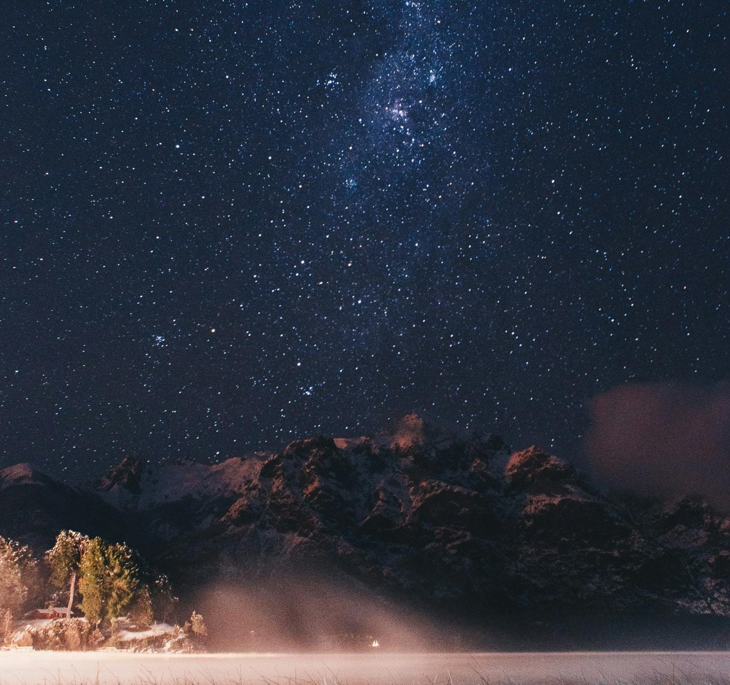 Starry sky with some trees and snowy mountains.
